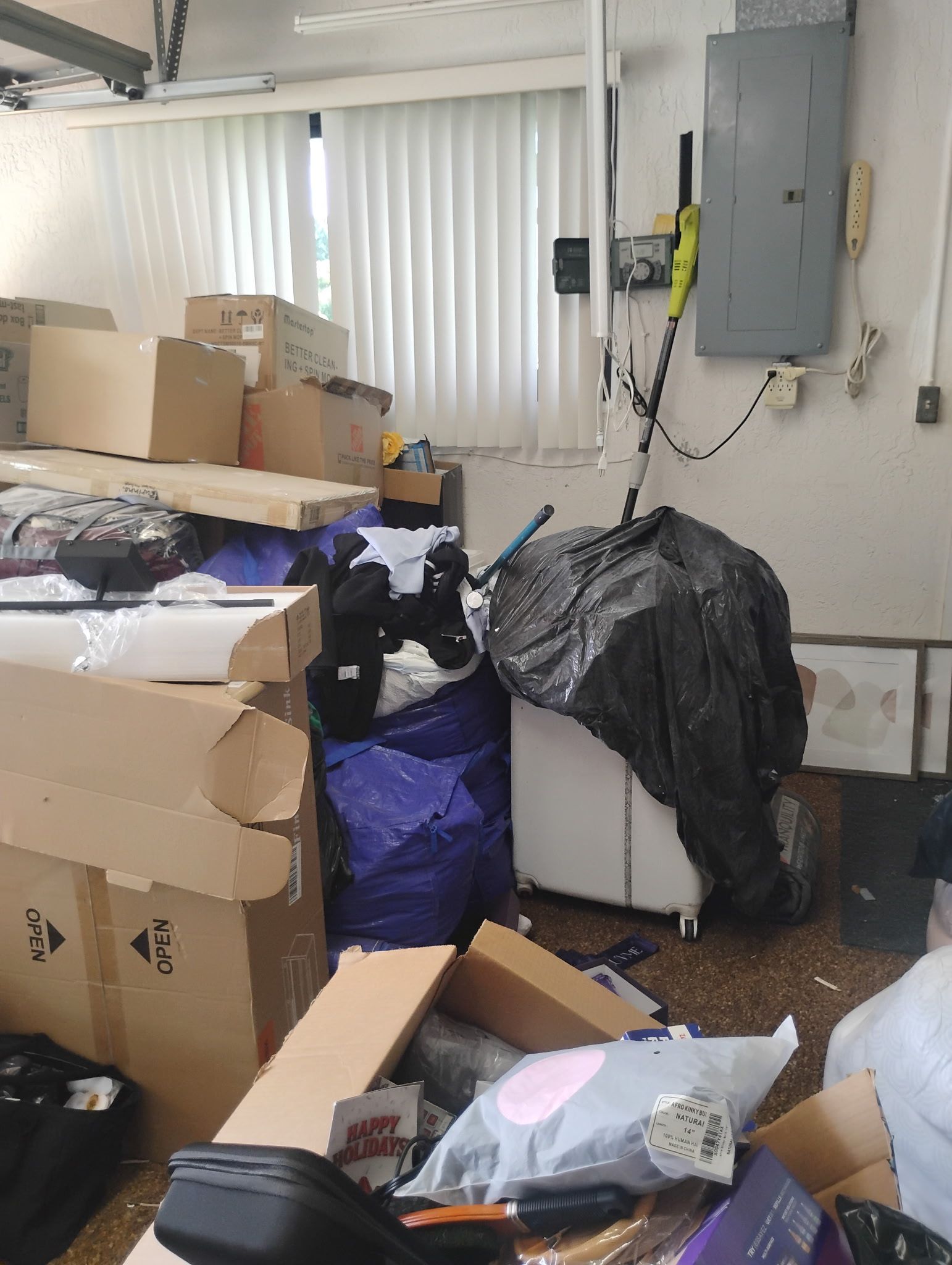 Cluttered garage with boxes, bags, and items strewn about. Gray electrical panel on the wall, partially visible window.