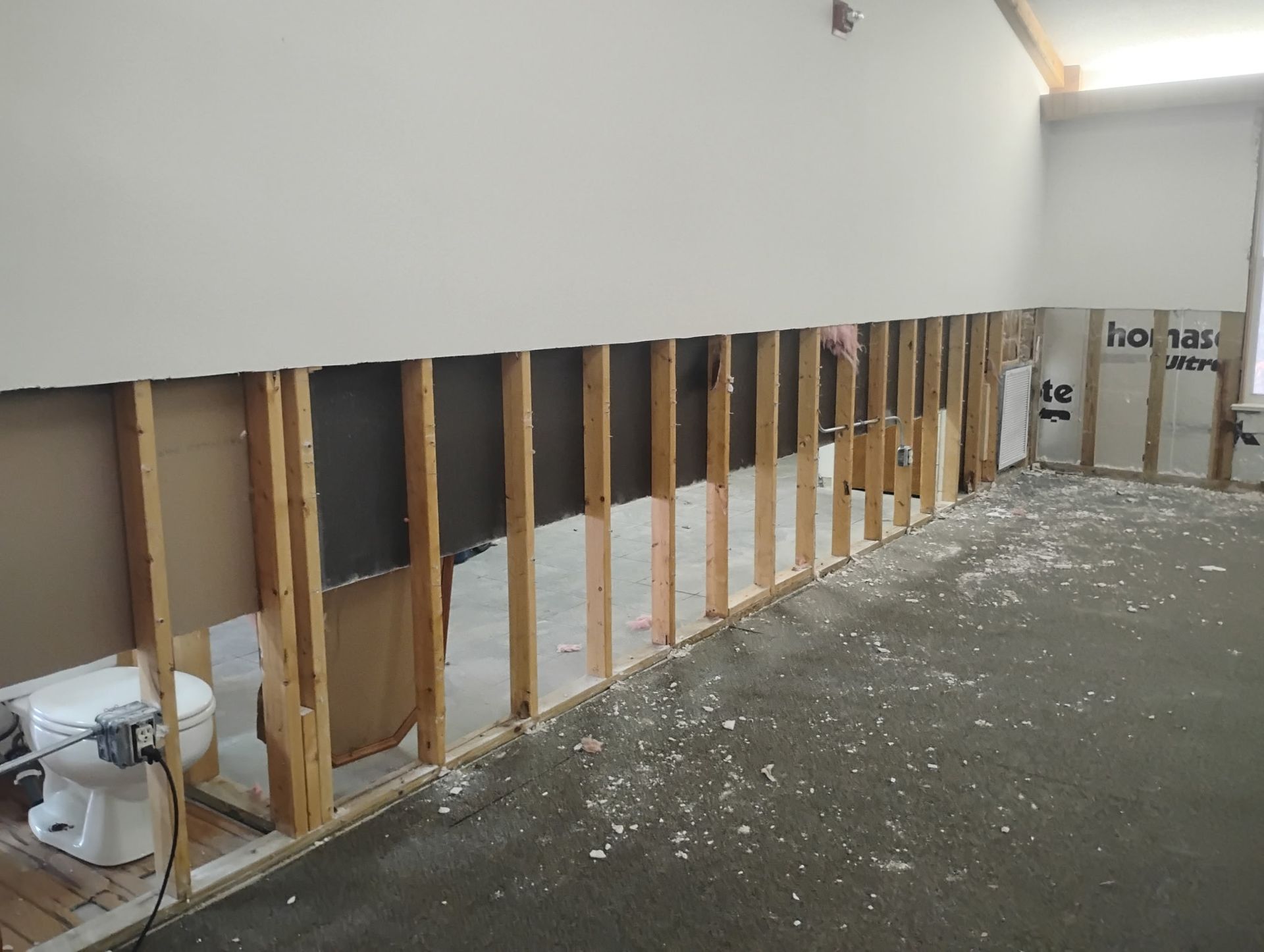 Wall of room partially demolished, revealing studs, insulation, and a toilet. Debris covers the floor.
