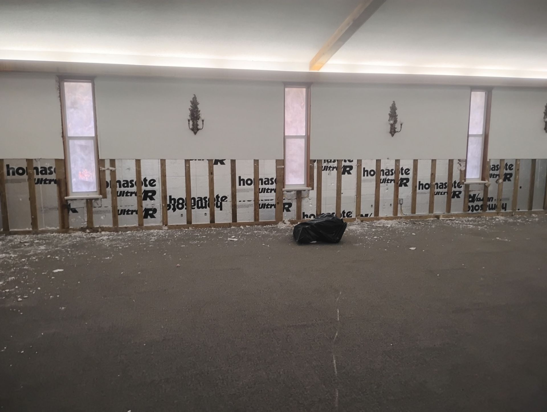 Interior view of a room under construction with exposed wood framing, three windows, and black insulation on the lower walls.
