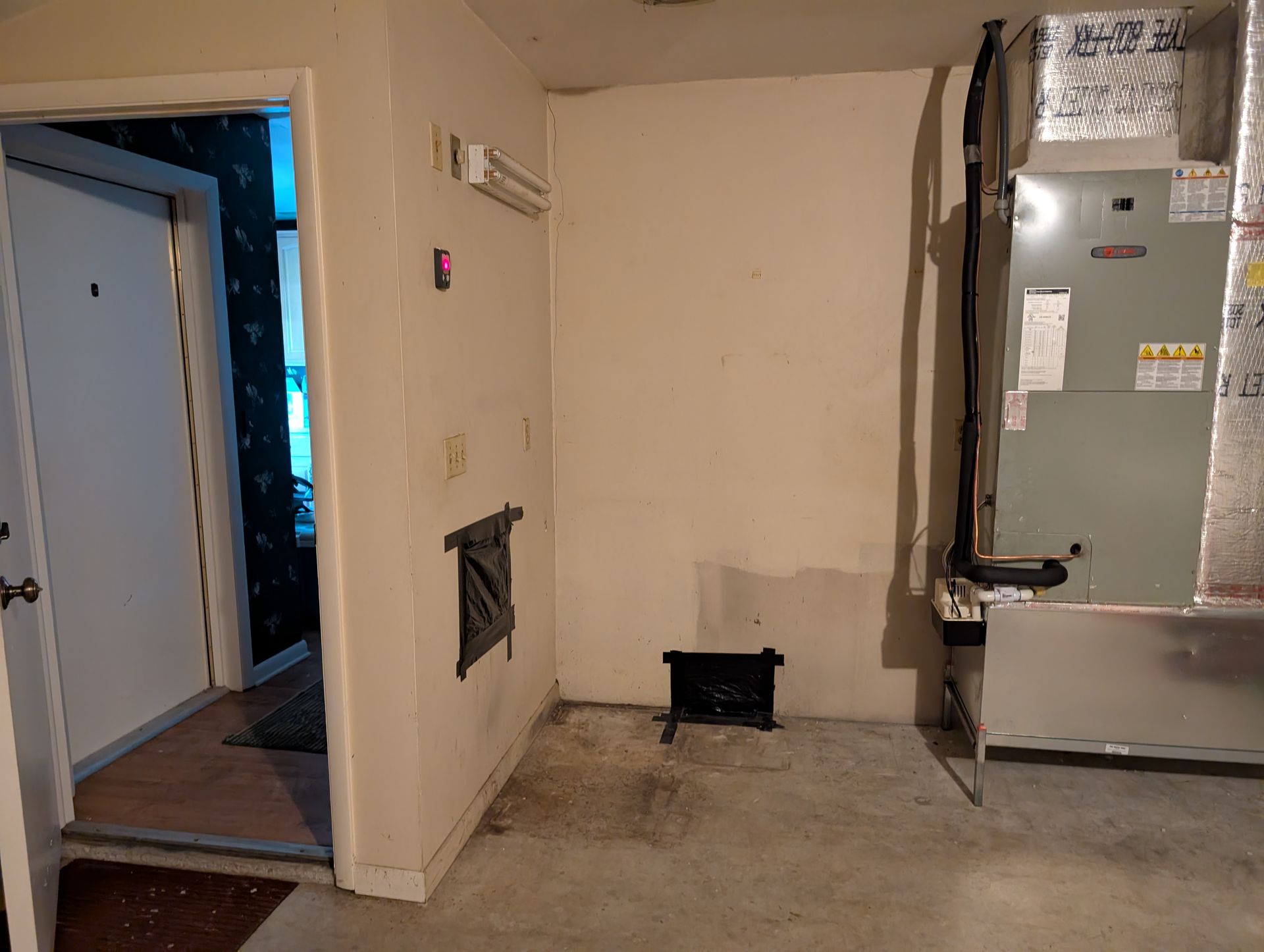 Interior room with HVAC unit, vent, door, and a small black object on the floor. Walls are off-white and floor is concrete.