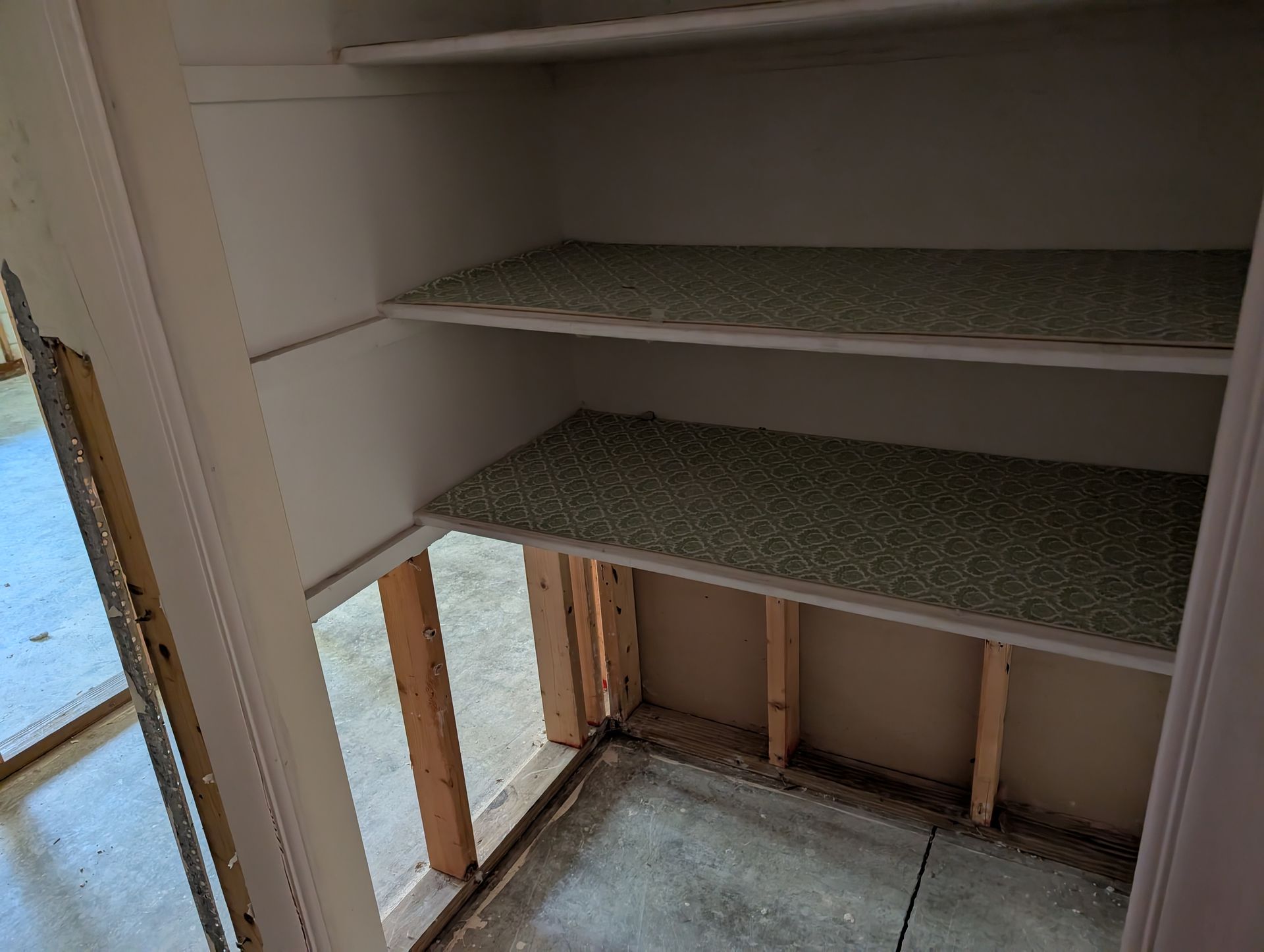 Empty closet with shelves, bare wooden framing and concrete floor. Shelves have textured covering.