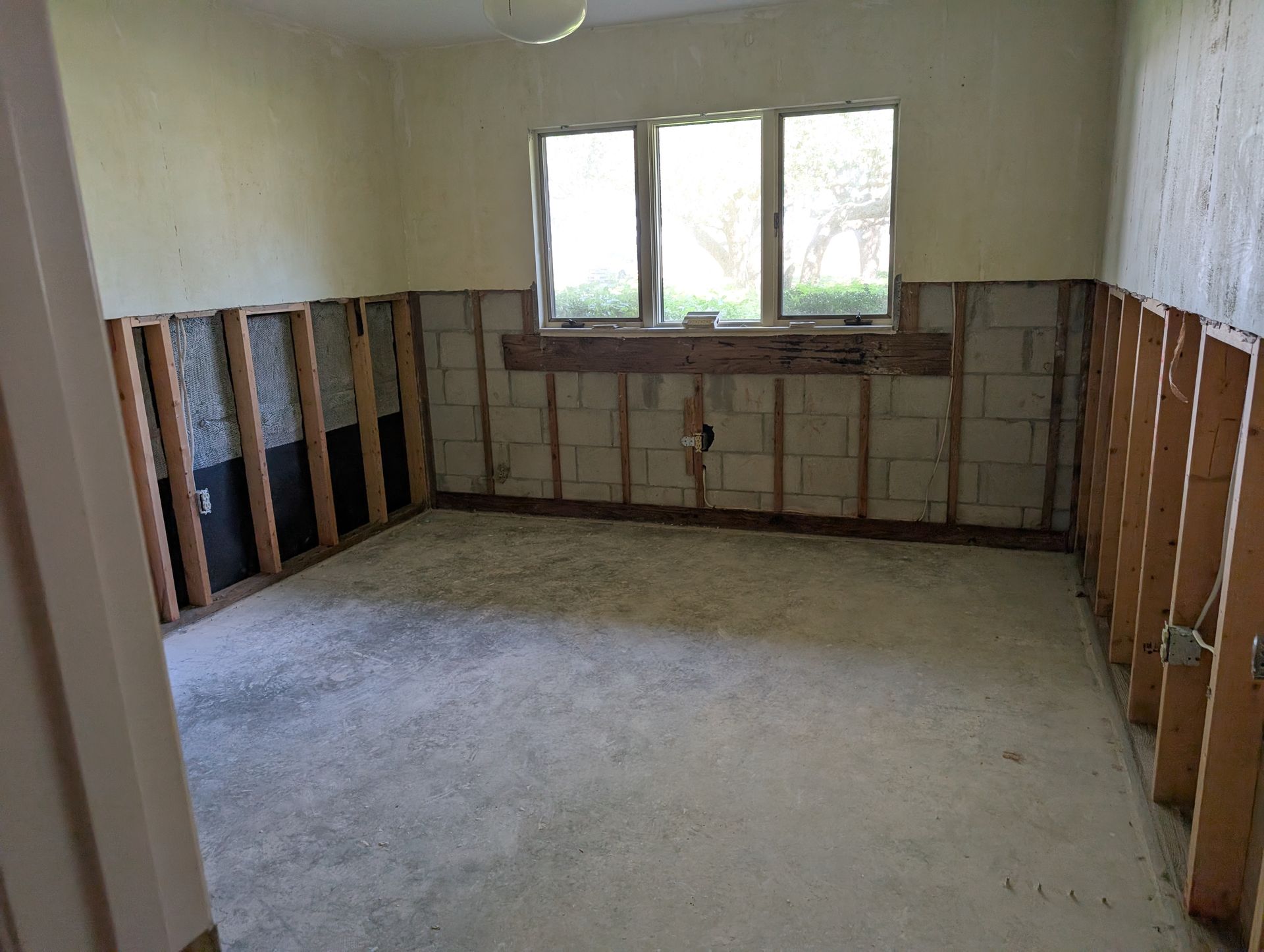 Room with exposed cinder block walls and studs, window, and concrete floor, appears under construction.