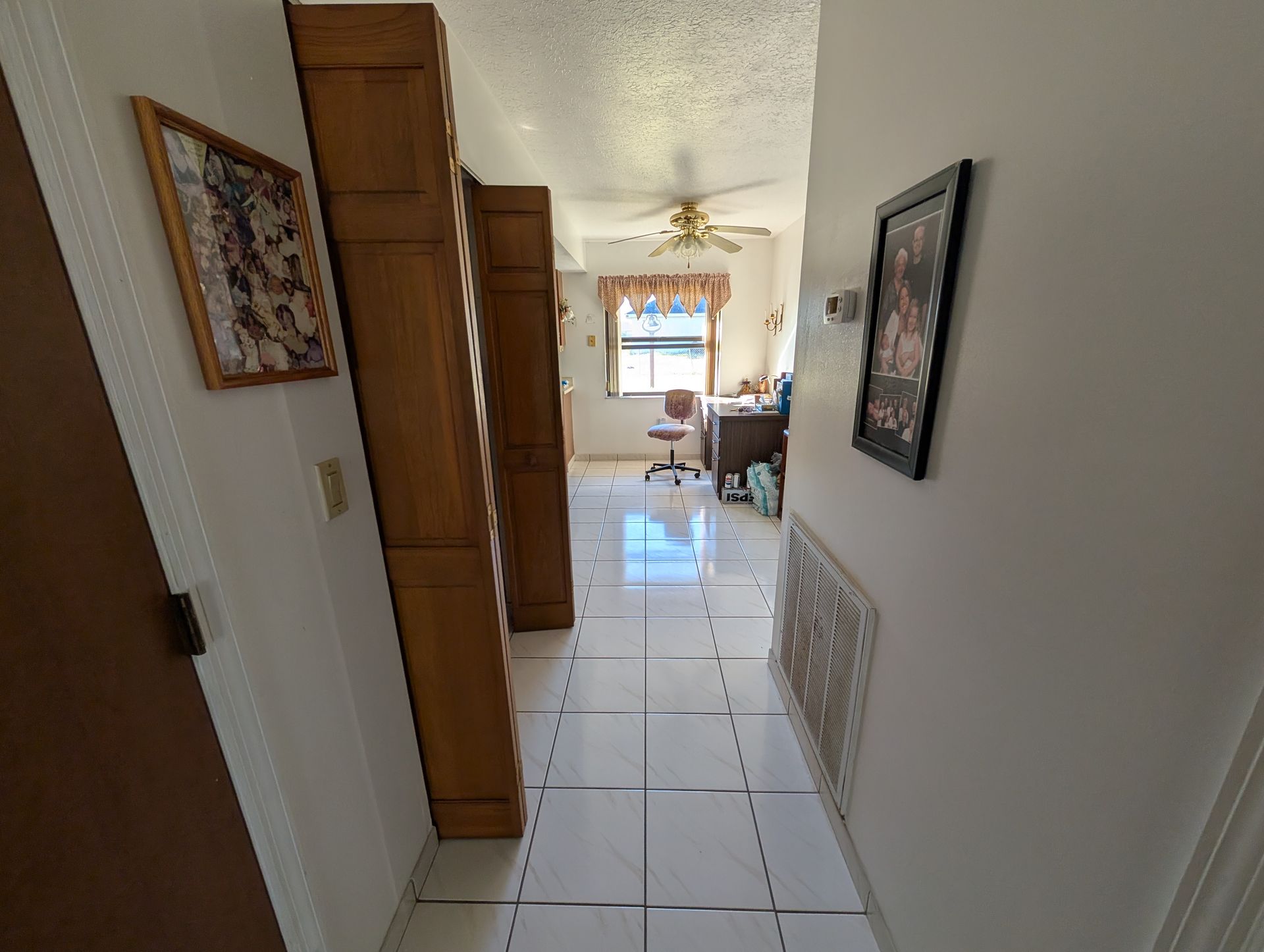 Hallway with tile floor leading to a room with a desk and window. Wooden shelves and artwork on walls.