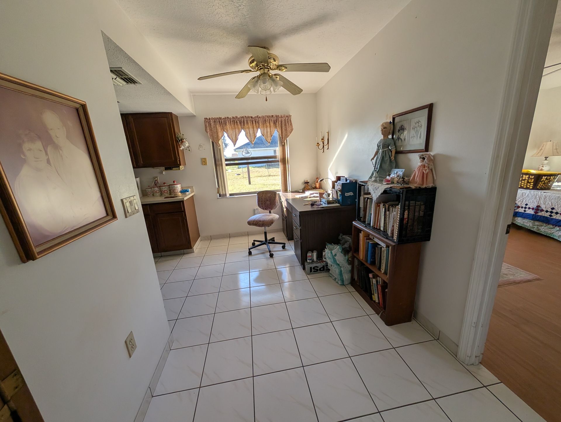 Small office space with desk, bookshelves, and kitchen visible. White tile floor and a window.
