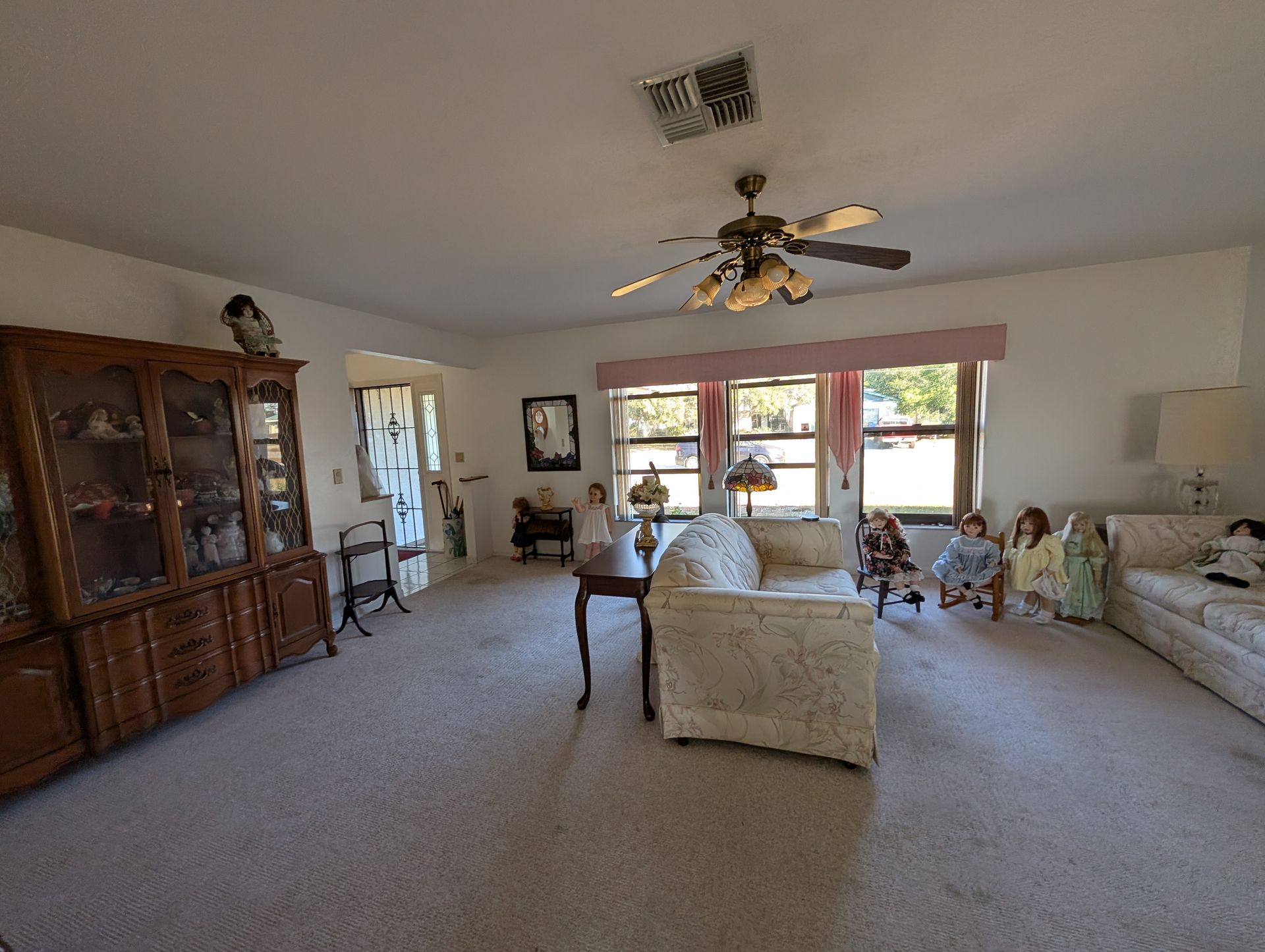 Living room with beige carpet, floral sofa, china cabinet, dolls, and ceiling fan.