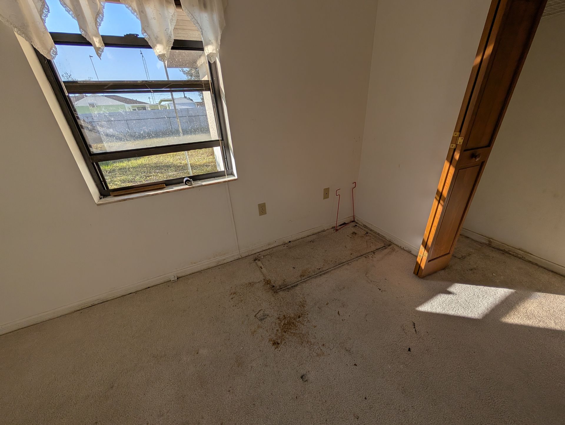 Small room with window, stained carpet, white walls, and a wooden beam leaning against the corner.