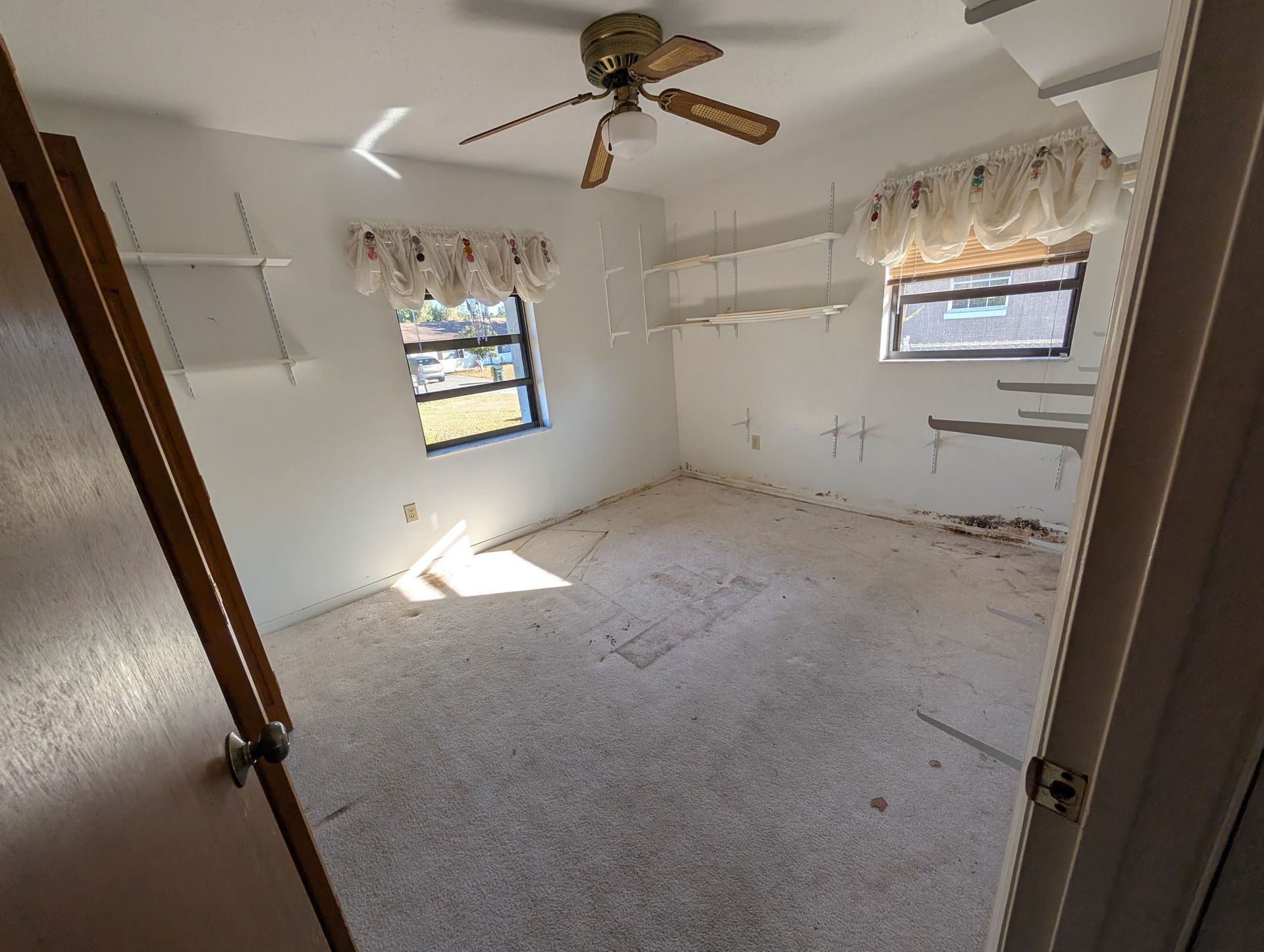 Empty room with a ceiling fan, two windows, shelves, and a partially open door.