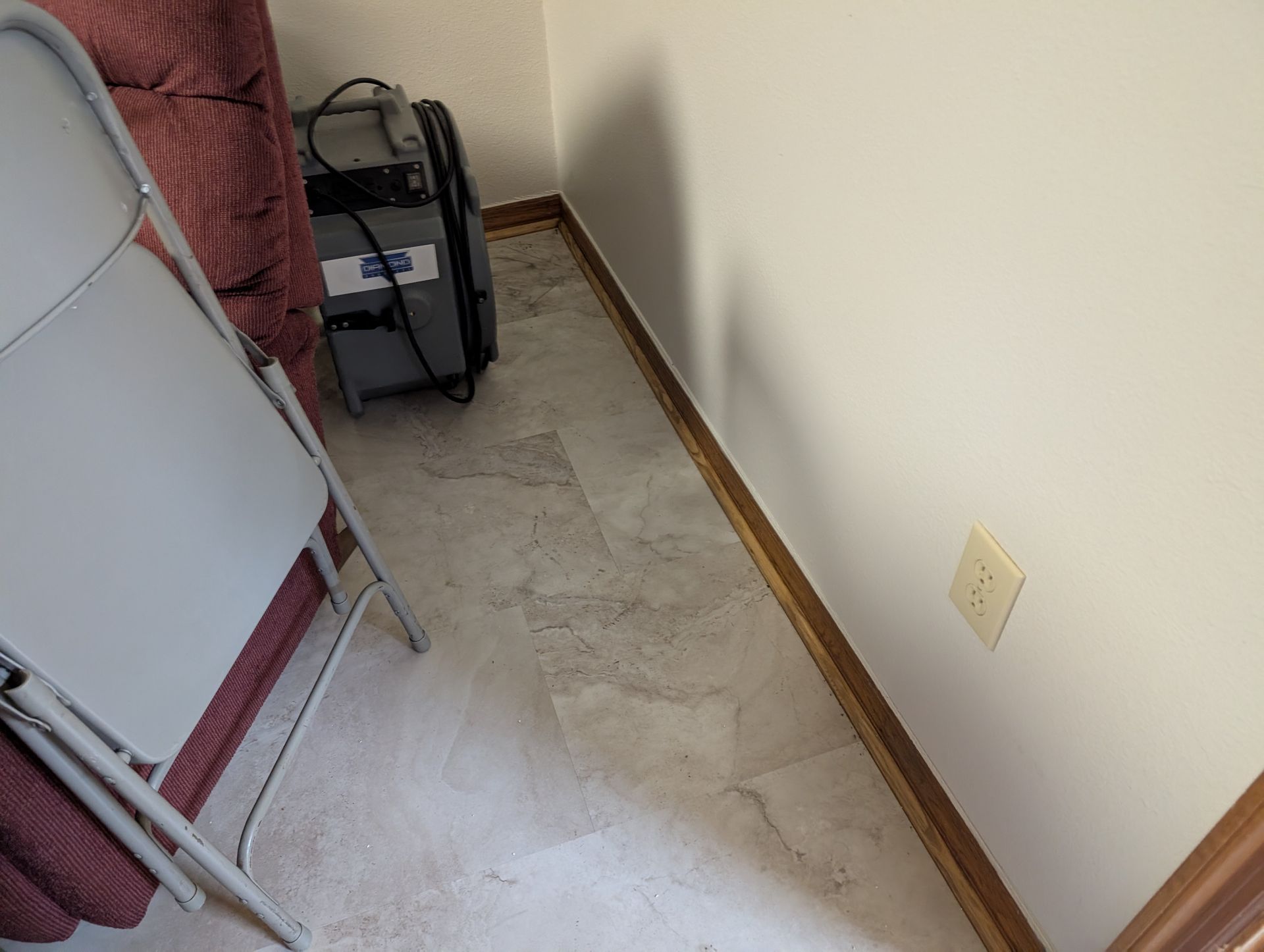 A corner room with tile floor, white walls, a folding chair, and a machine.