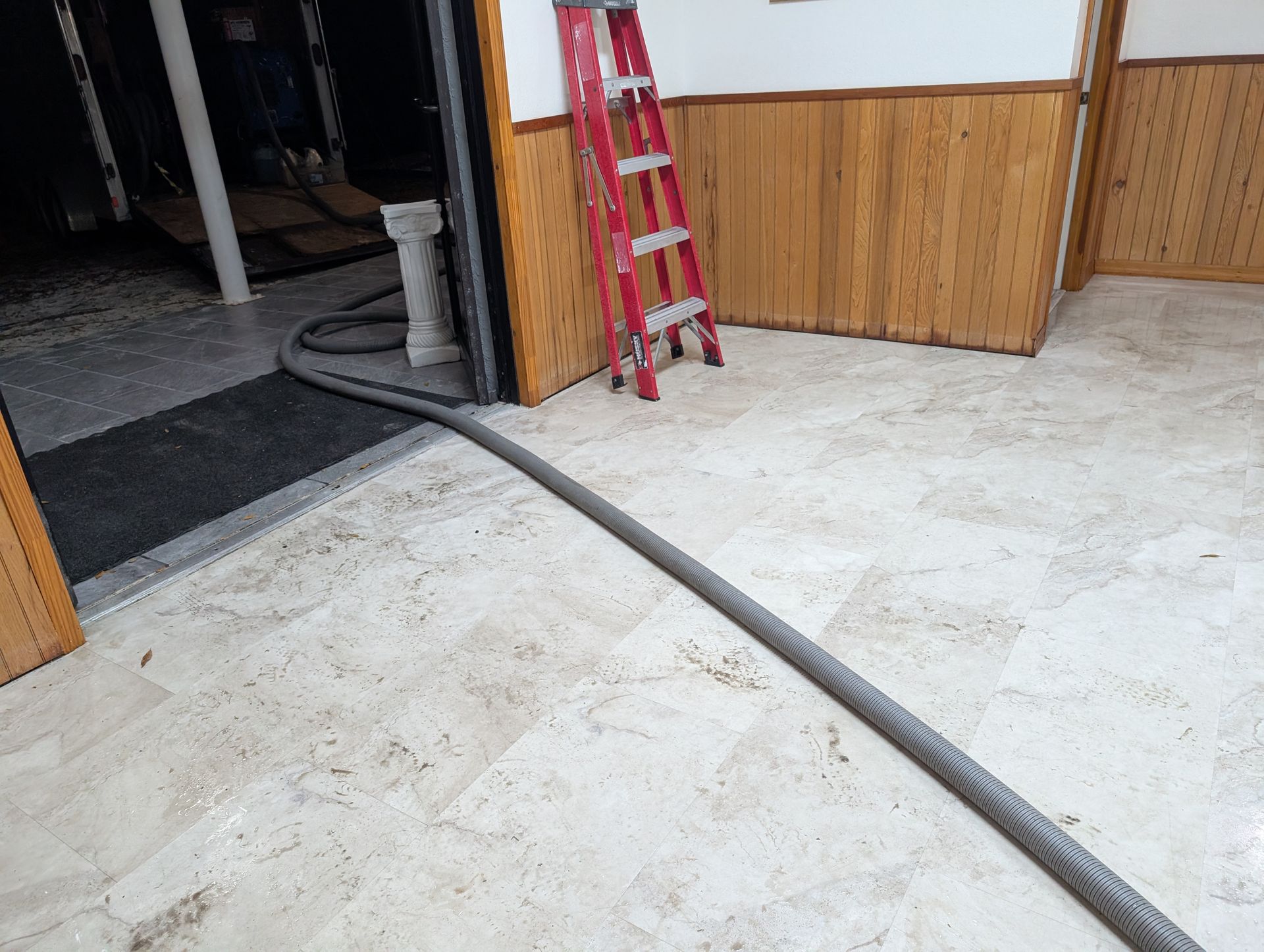 A gray hose extends across a tiled floor toward a doorway with a red ladder and wood paneling visible.