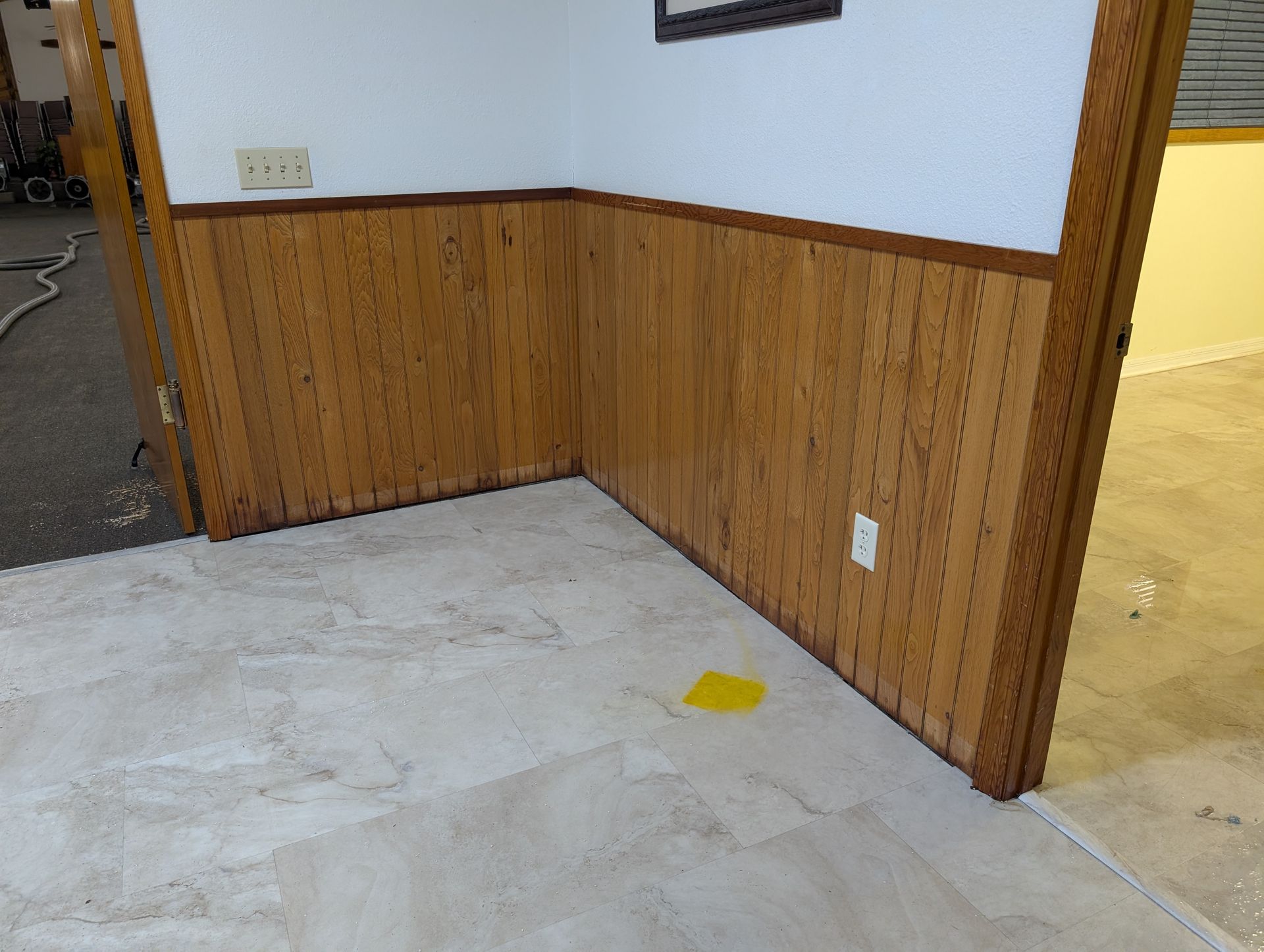 Corner of a room with wood paneling and beige tile flooring, a yellow sticky note is on the floor.