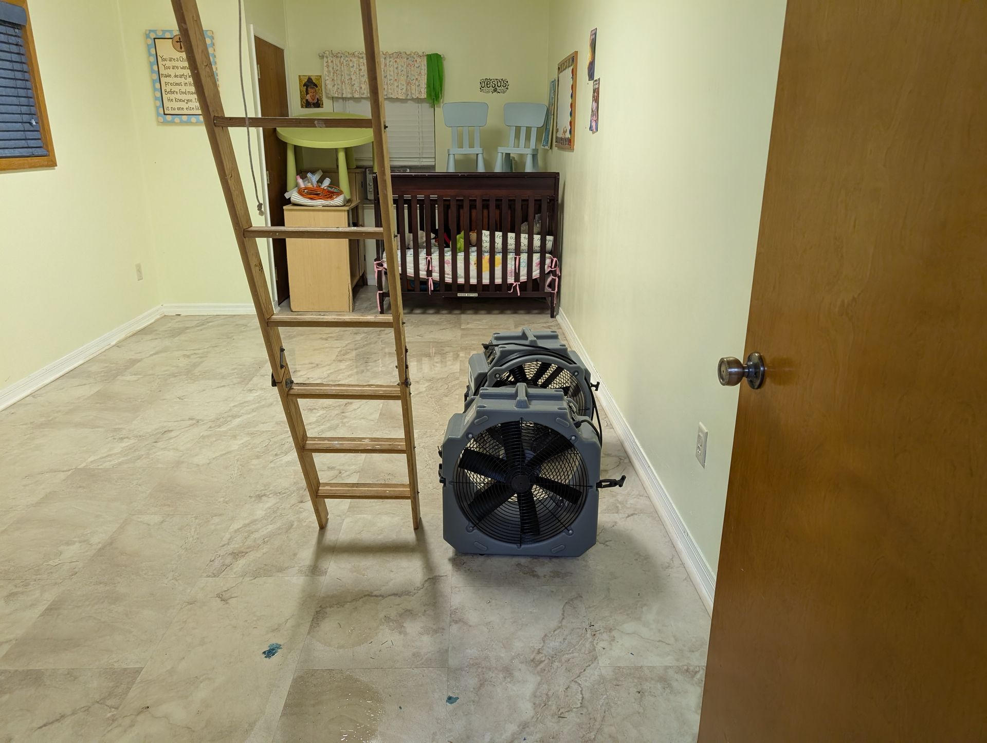 Room with a ladder, large gray fan, and a crib. Light yellow walls, wooden door.