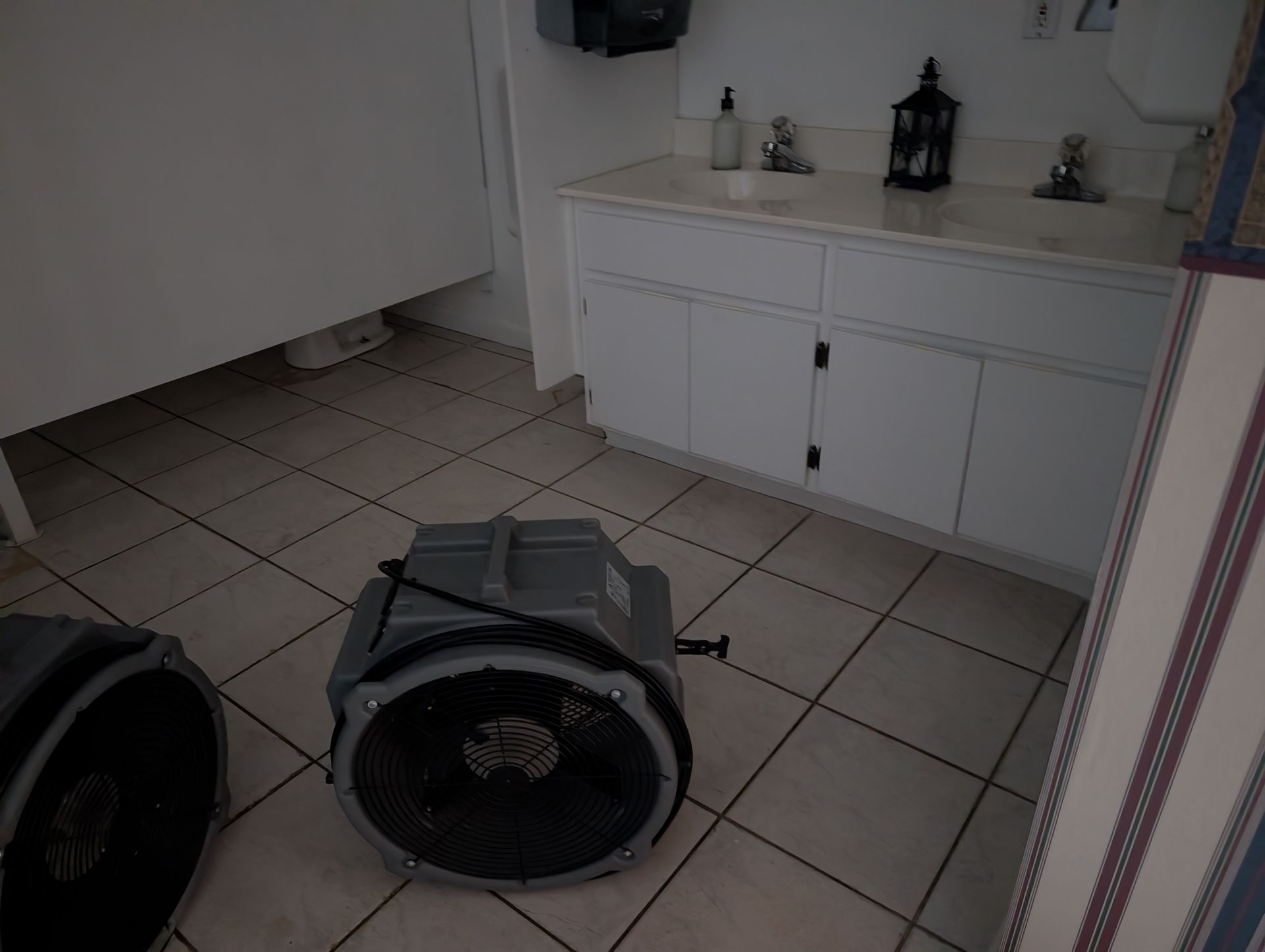 Two industrial fans drying a tiled bathroom floor. White cabinets, sinks, and a stall are visible.