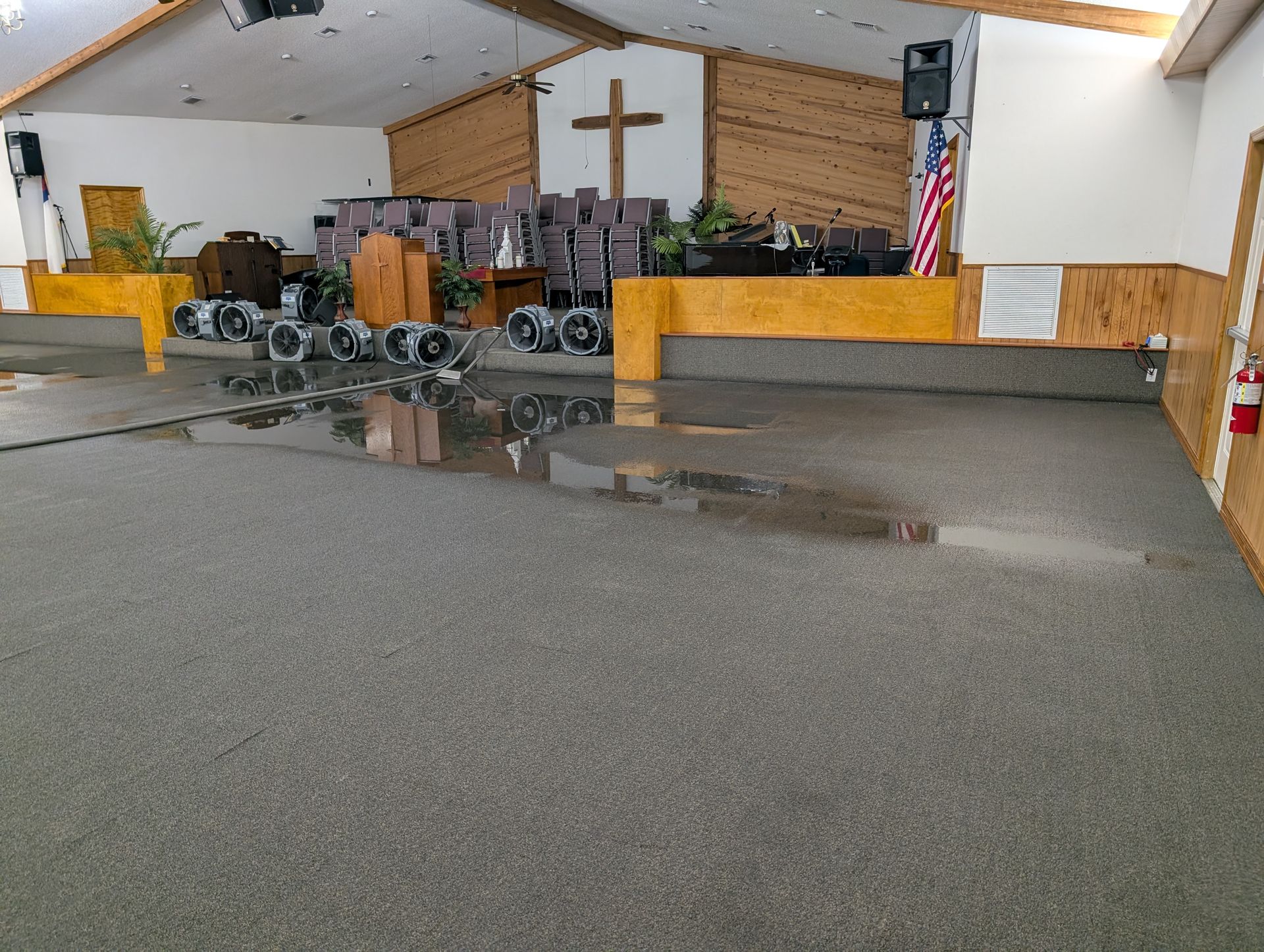 Church interior with water damage on the carpet, industrial fans running; cross, American flag in background.