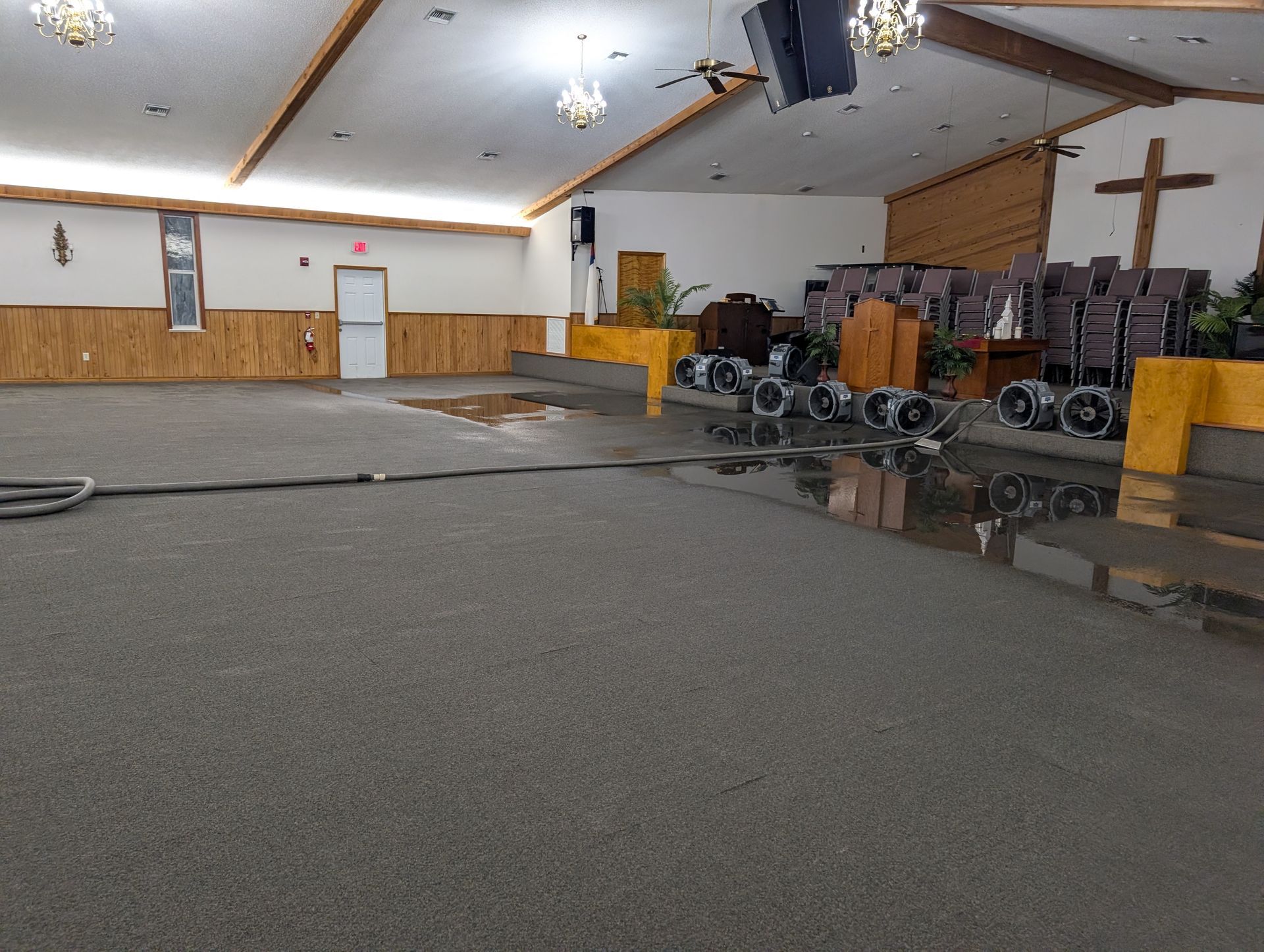 Flooded church interior with fans drying the carpet. Brown cross, pews, and exit door visible.