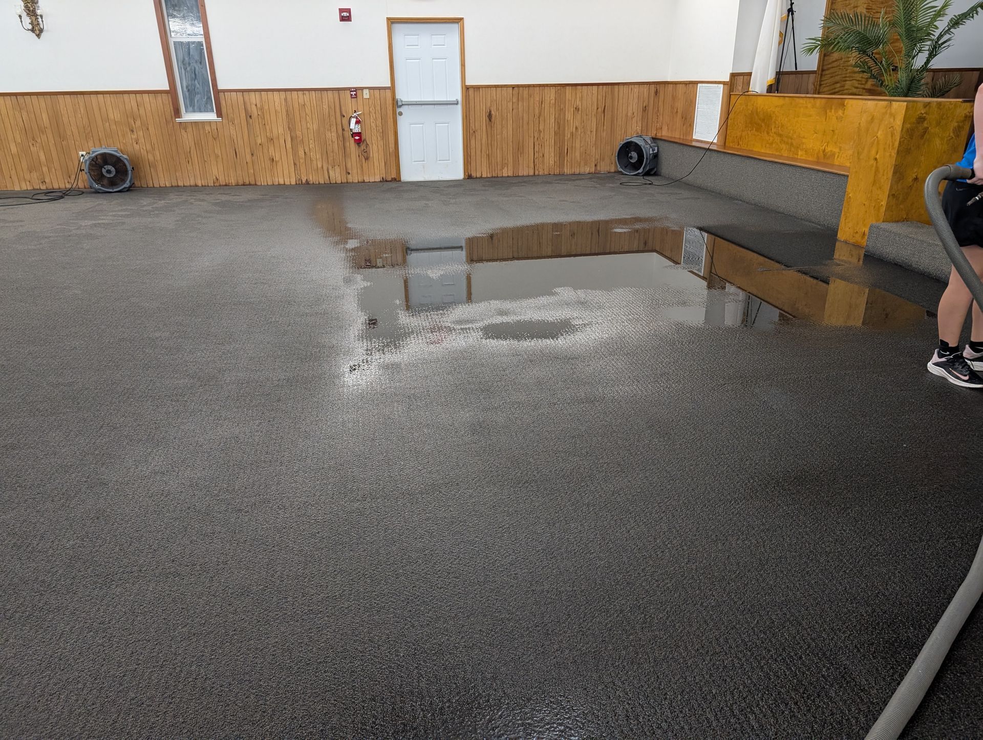Indoor space with standing water on a dark gray floor, fans blowing, person with hose on the right, wood panel walls.