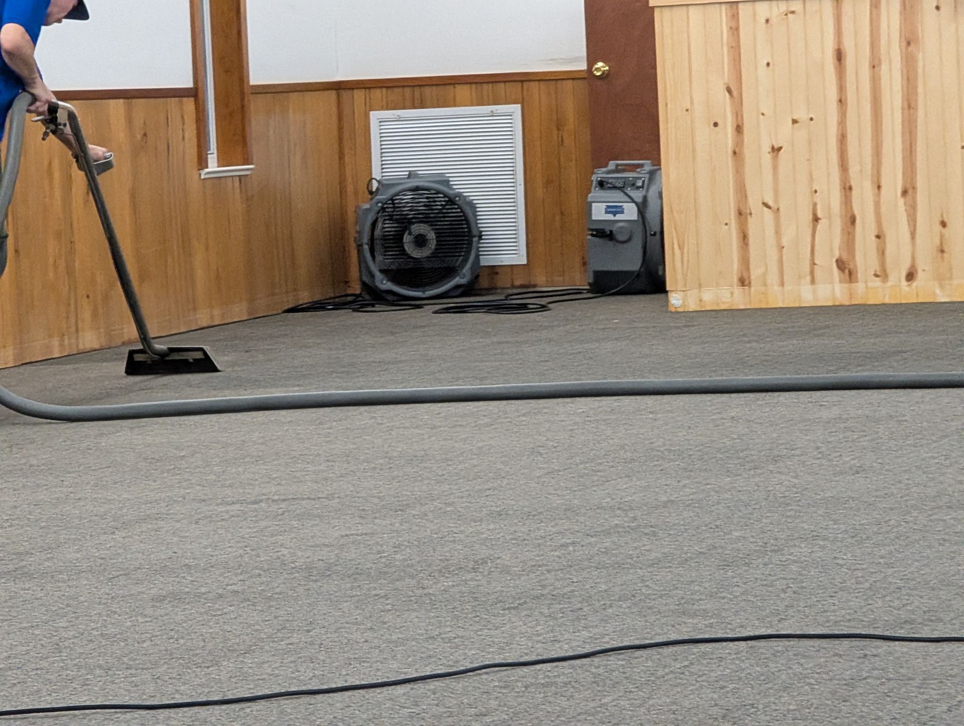 Person vacuuming carpeted room with equipment, fan, and paneling.