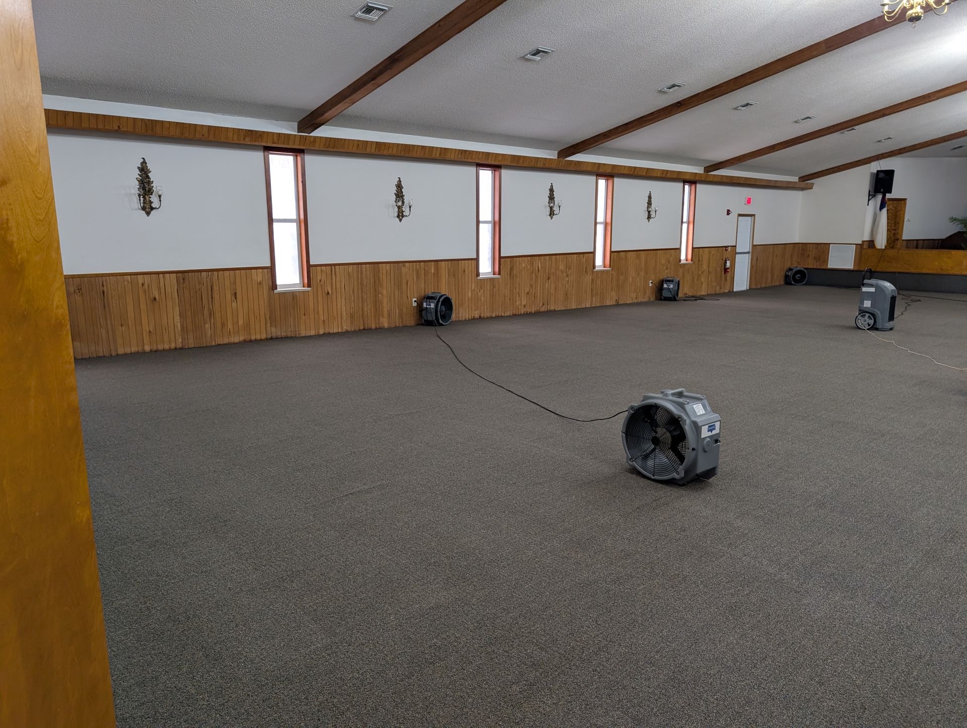 Empty event space with carpet and several fans on the floor; windows along the wall; wood trim.
