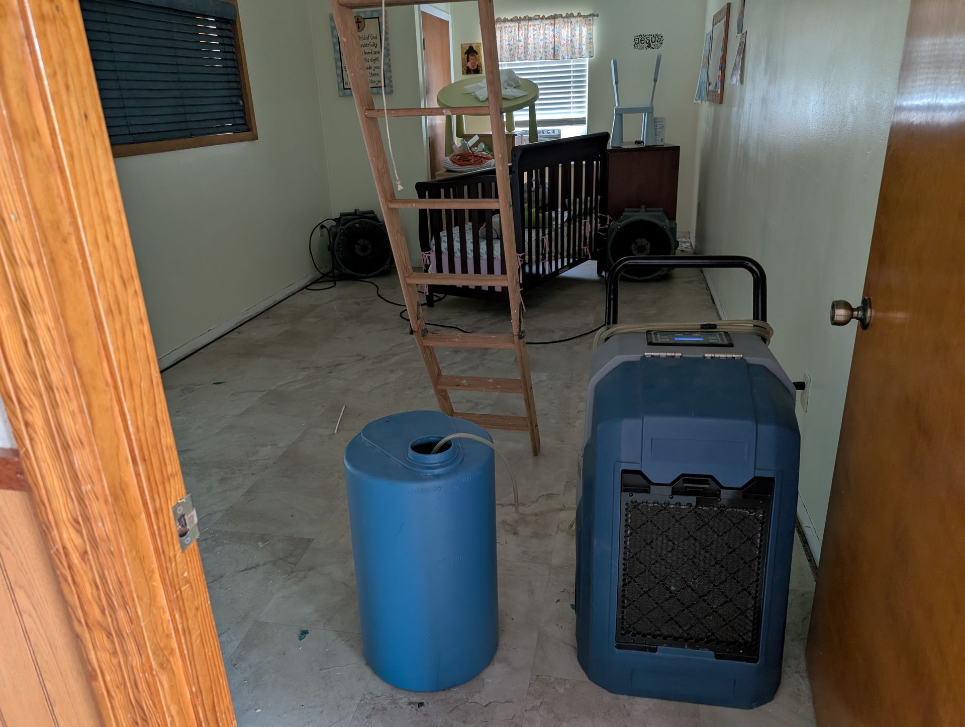 Room with a dehumidifier, blue barrel, crib, and ladder; door open.