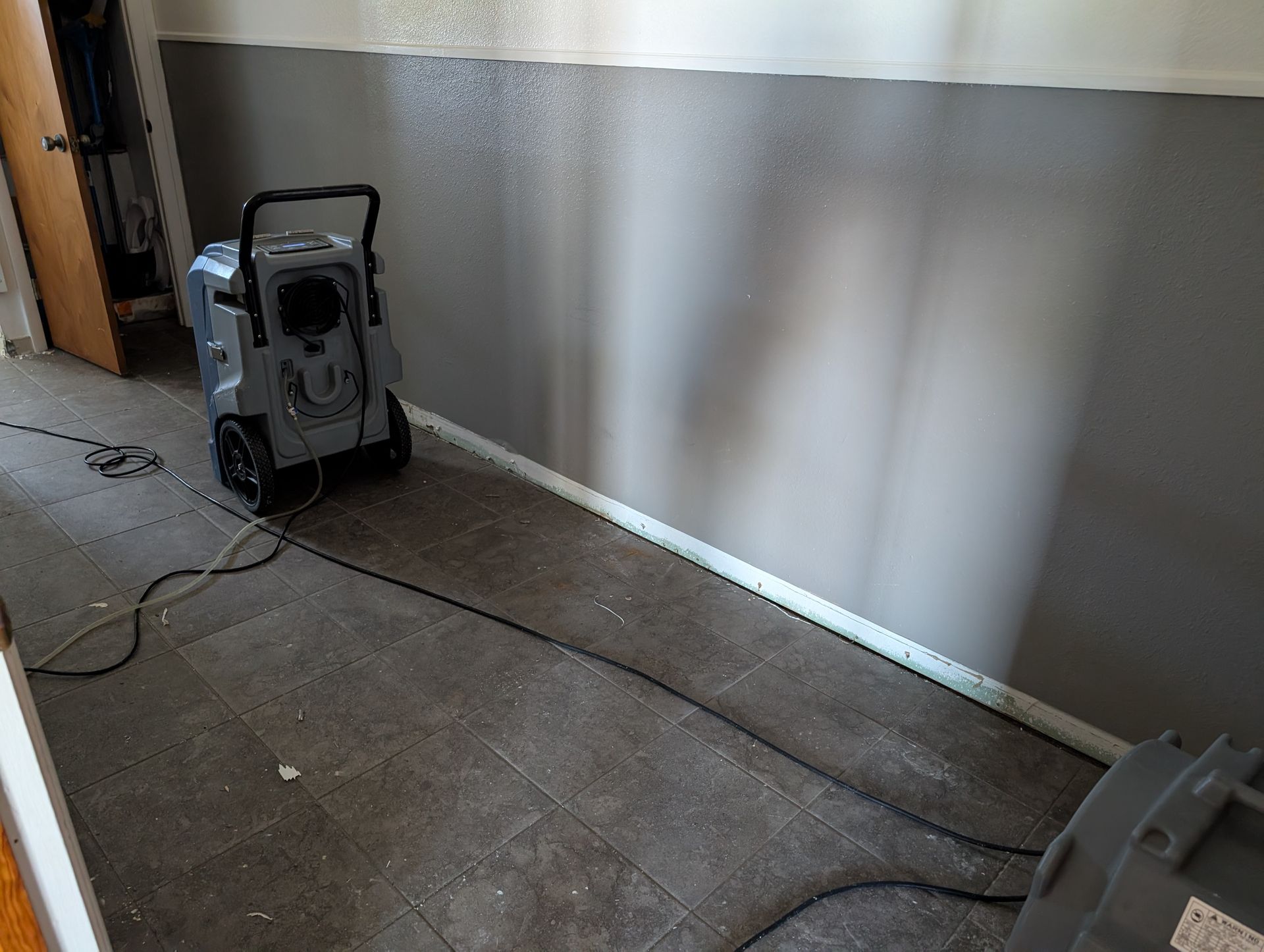 Dehumidifier on tiled floor next to a wall painted gray and white trim. Electrical cord is visible.