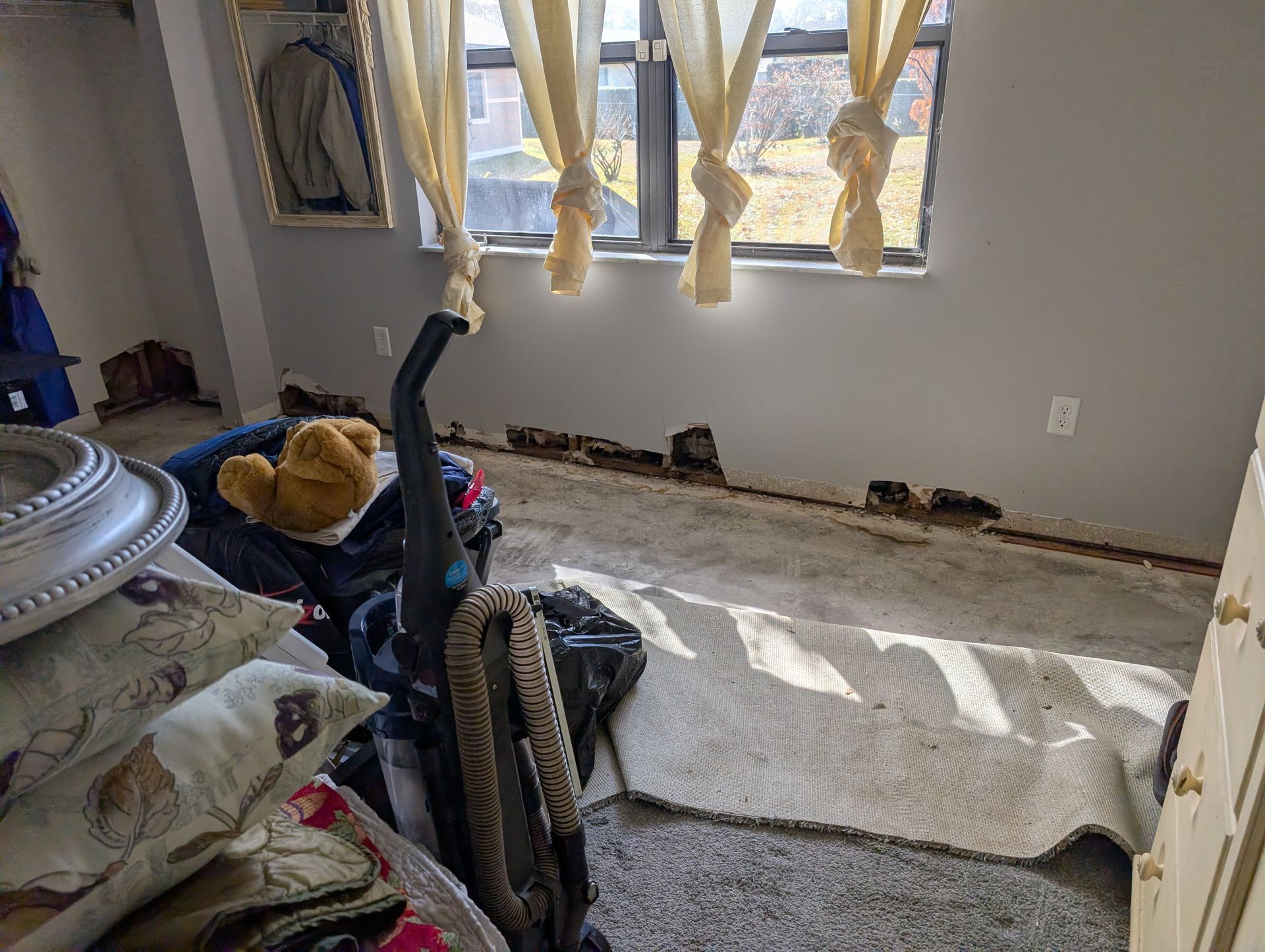 Room with damaged walls, debris, vacuum, and curtains in front of a window.