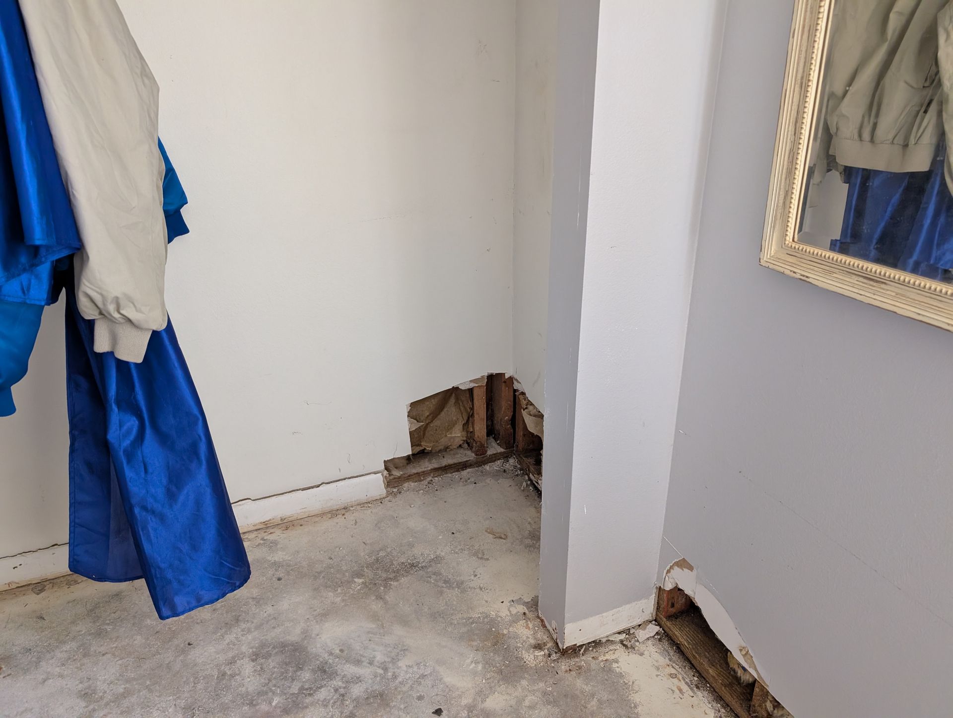 Damaged wall corner, showing exposed wood and crumbling plaster. Clothes hang nearby, mirror visible.