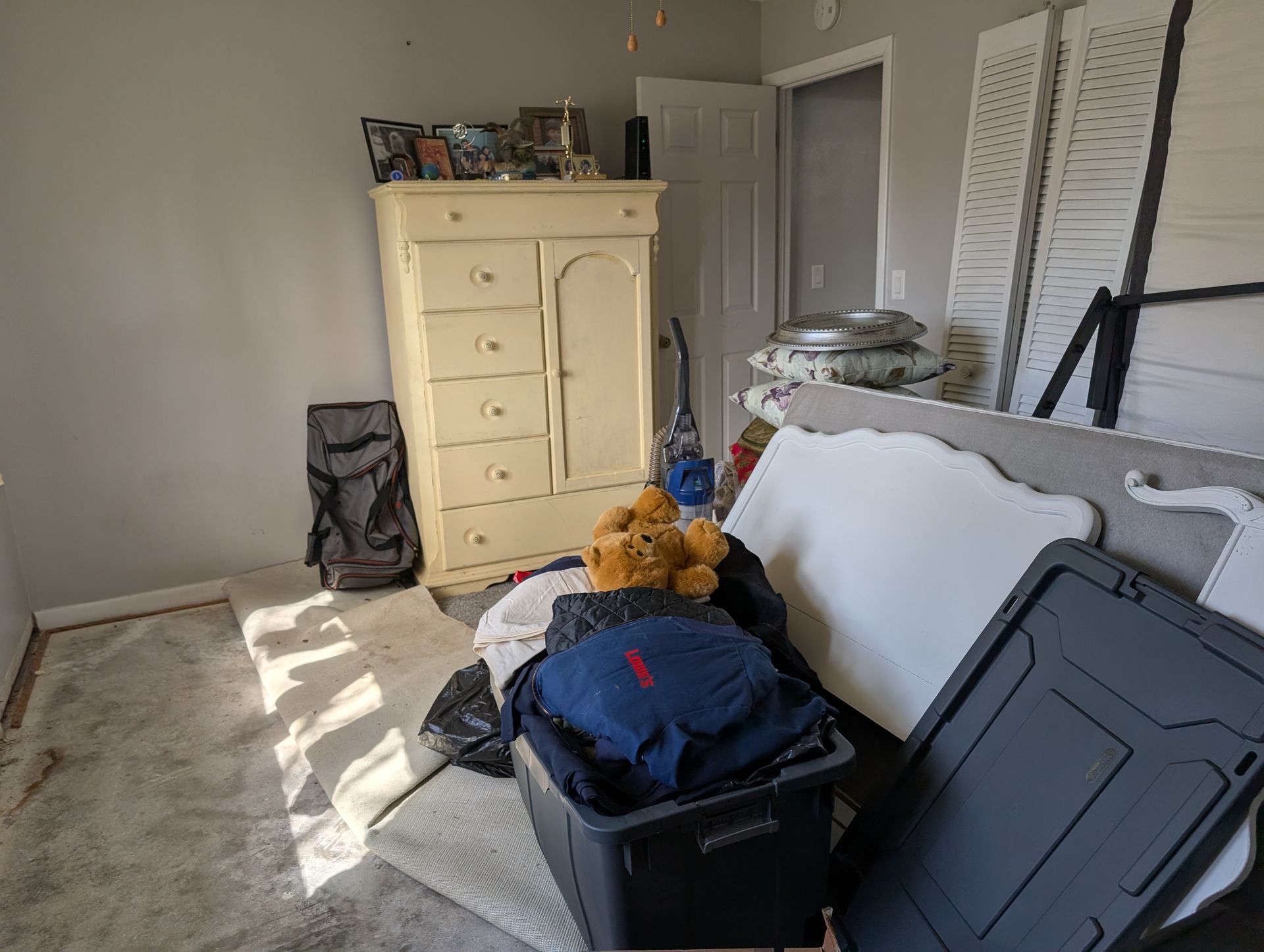 A cluttered bedroom with a light-colored dresser, storage bin, and items on the floor.