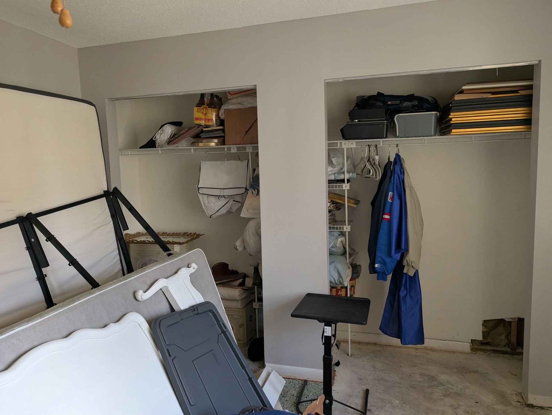Two closets with shelving, one with clothes hanging, cluttered with items and a bed frame.