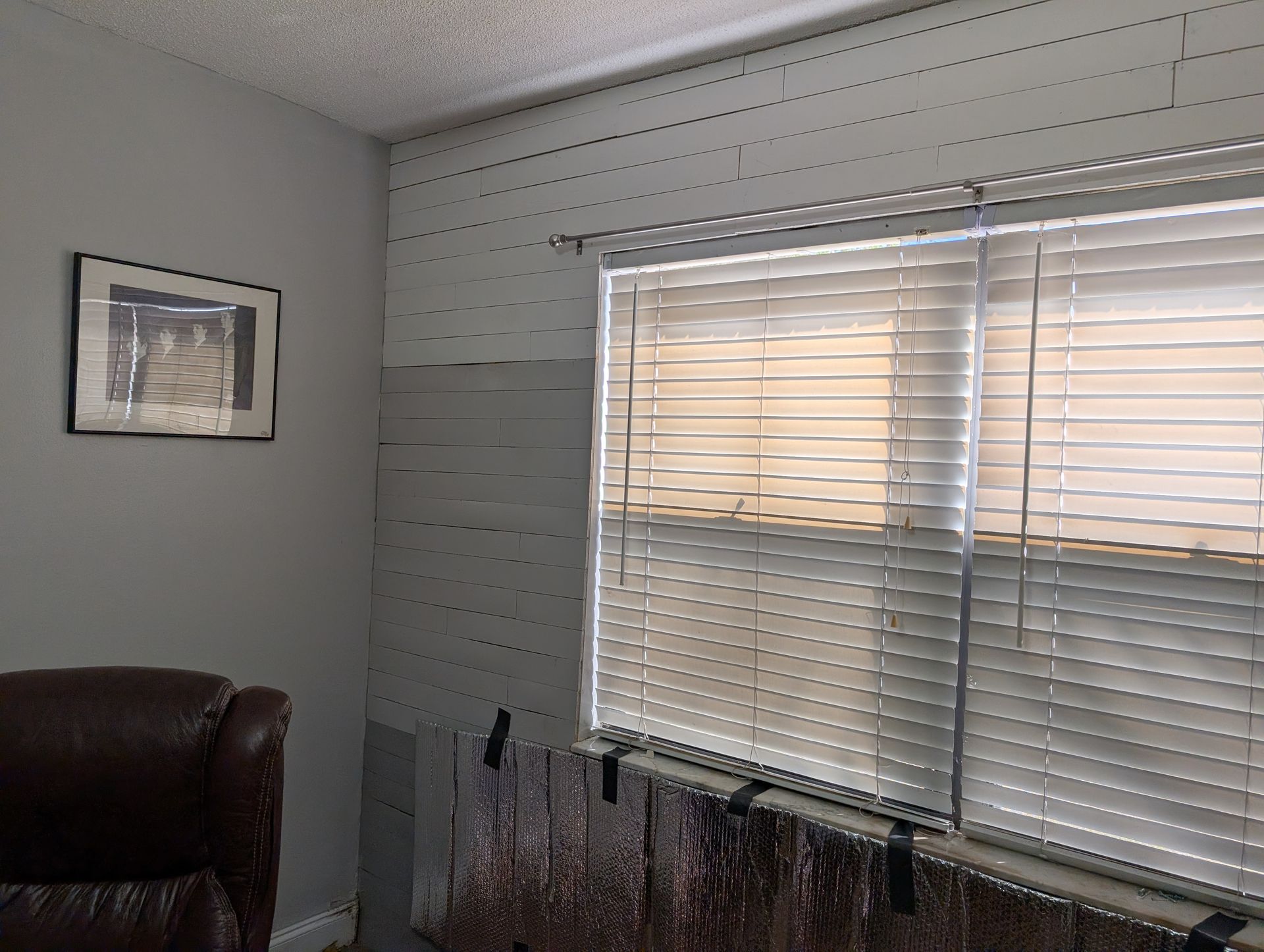 Room with a window, blinds, art, and partially covered wall; gray walls, brown couch.