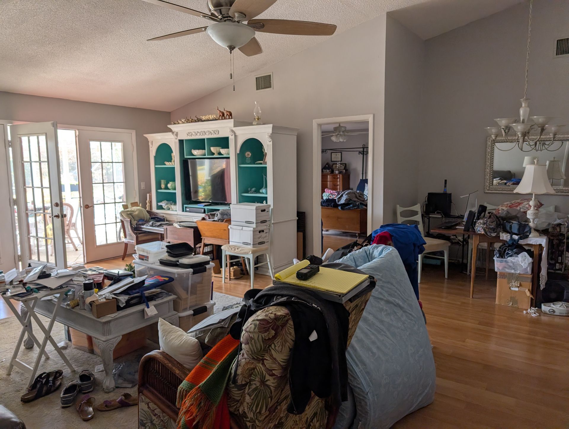 Cluttered living room with various items, including furniture, clothing, and papers. Hardwood floors, natural light.