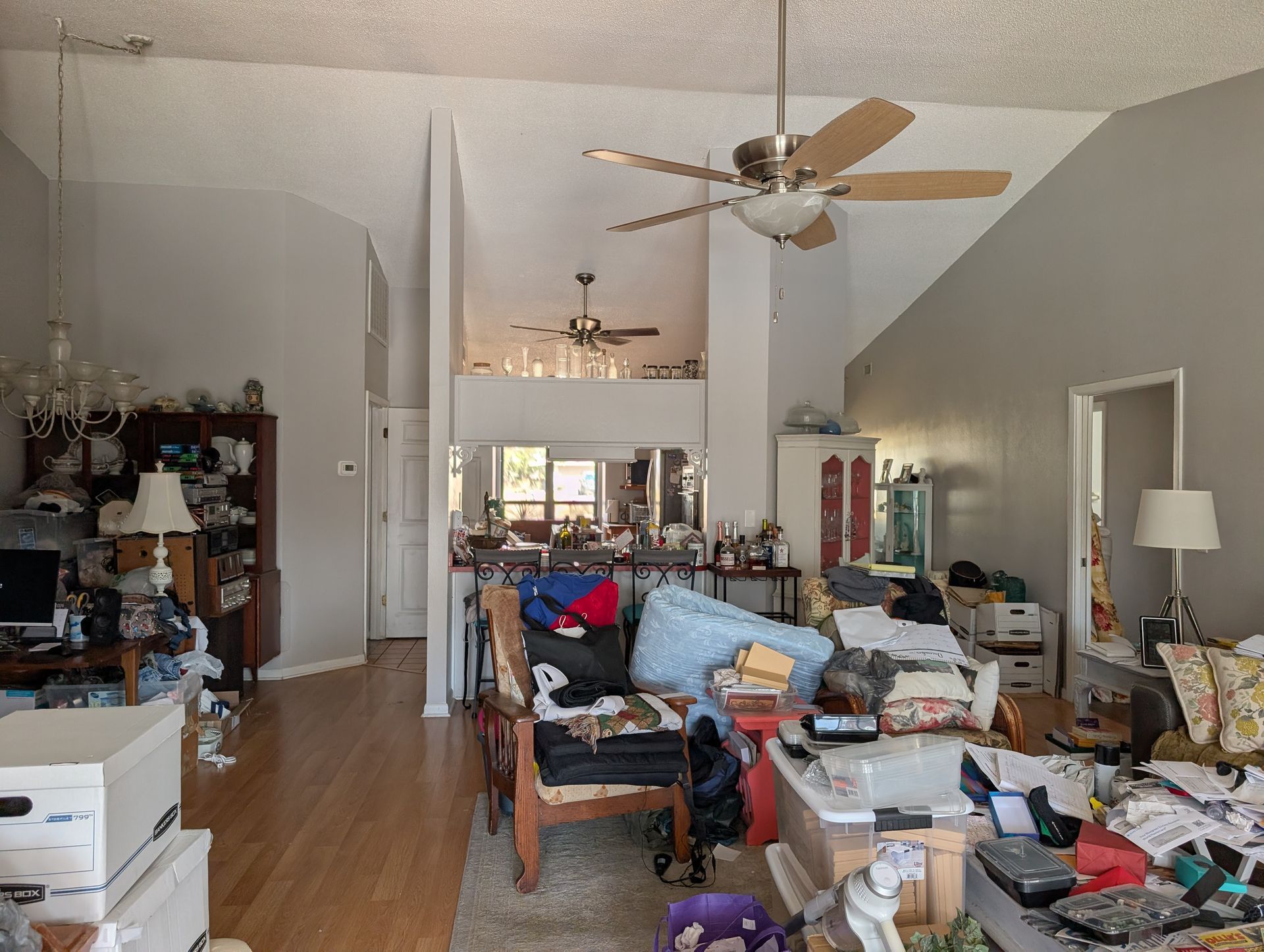 Cluttered living room with high ceilings, lots of stuff piled on furniture and floor.