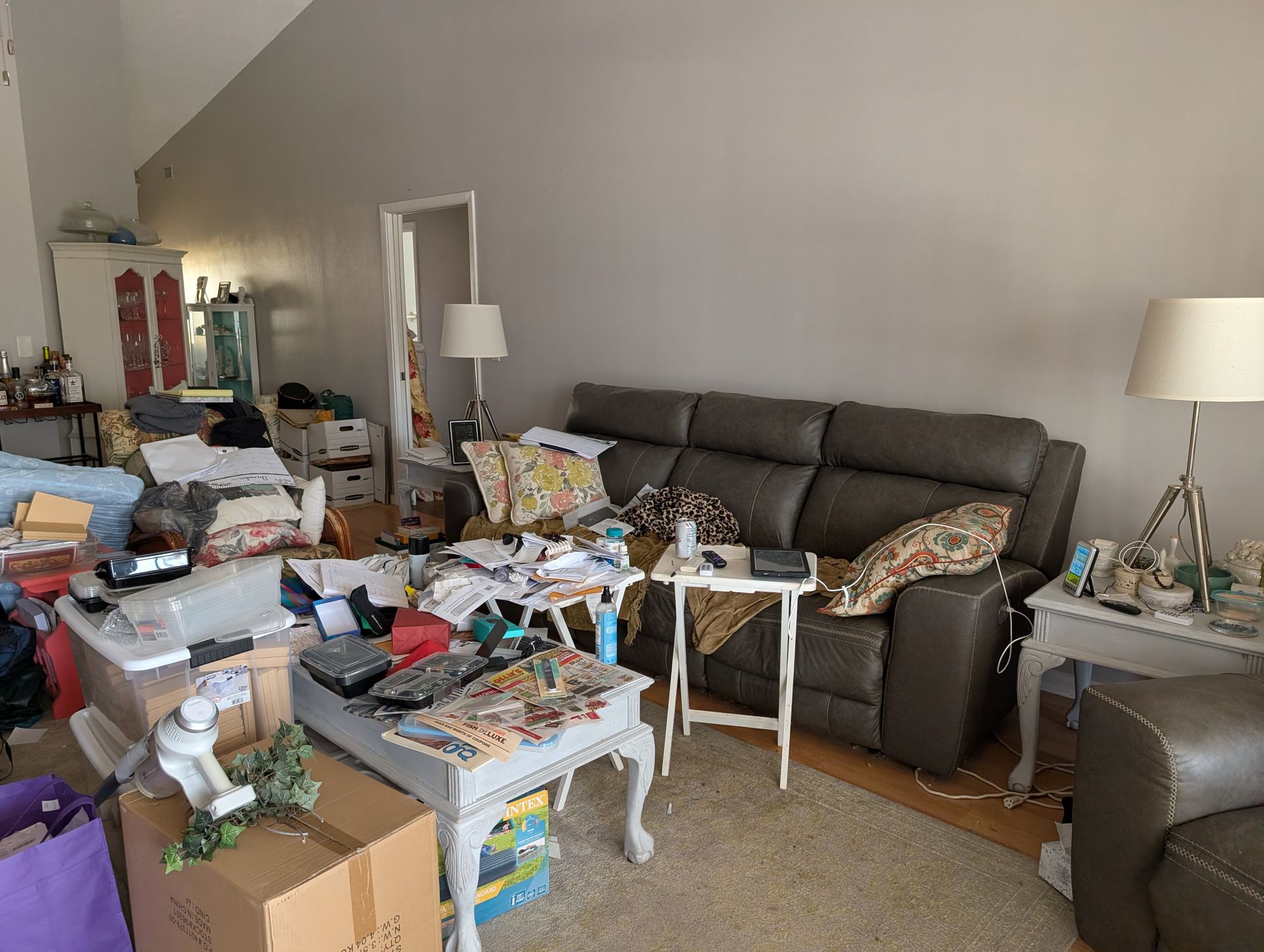 Cluttered living room with a gray recliner, two end tables, and boxes of items.