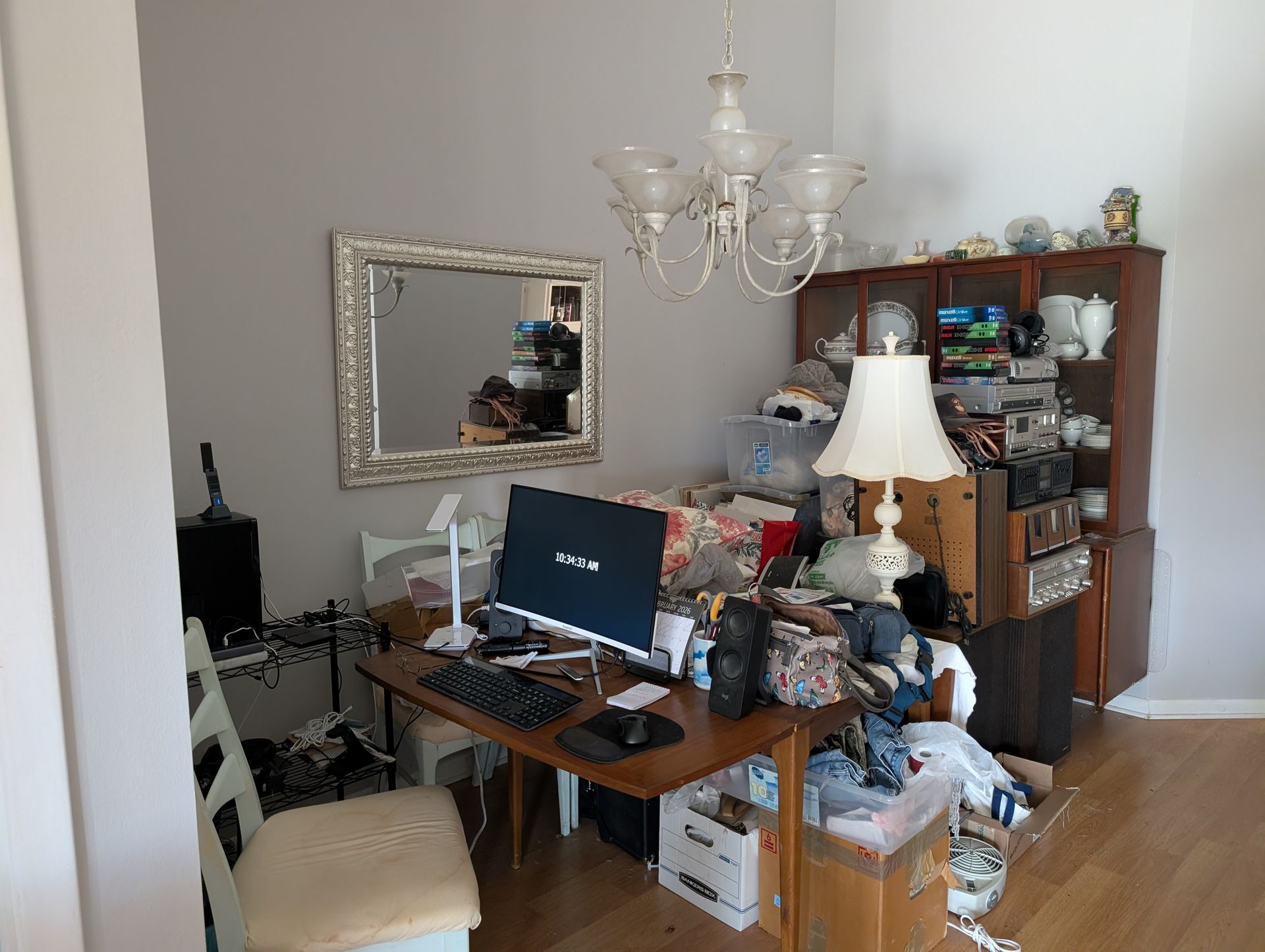A cluttered home office with a computer desk, shelving unit, and a chair. A mirror and chandelier are also present.