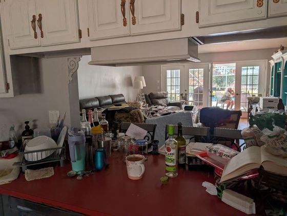 Messy kitchen counter with clutter. Living room visible through the open doorway and a person sitting outside.