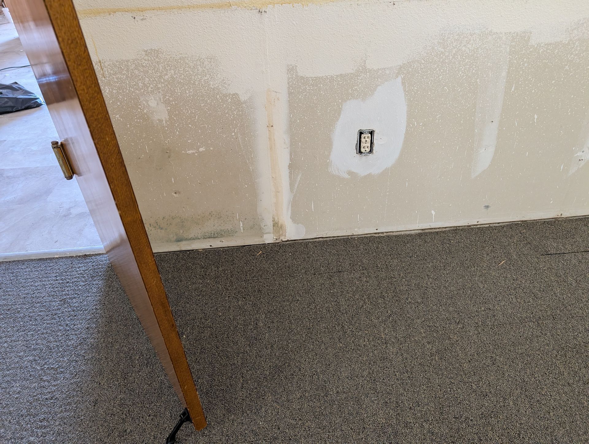 A partially exposed wall with an electrical outlet, next to a cabinet on carpet.