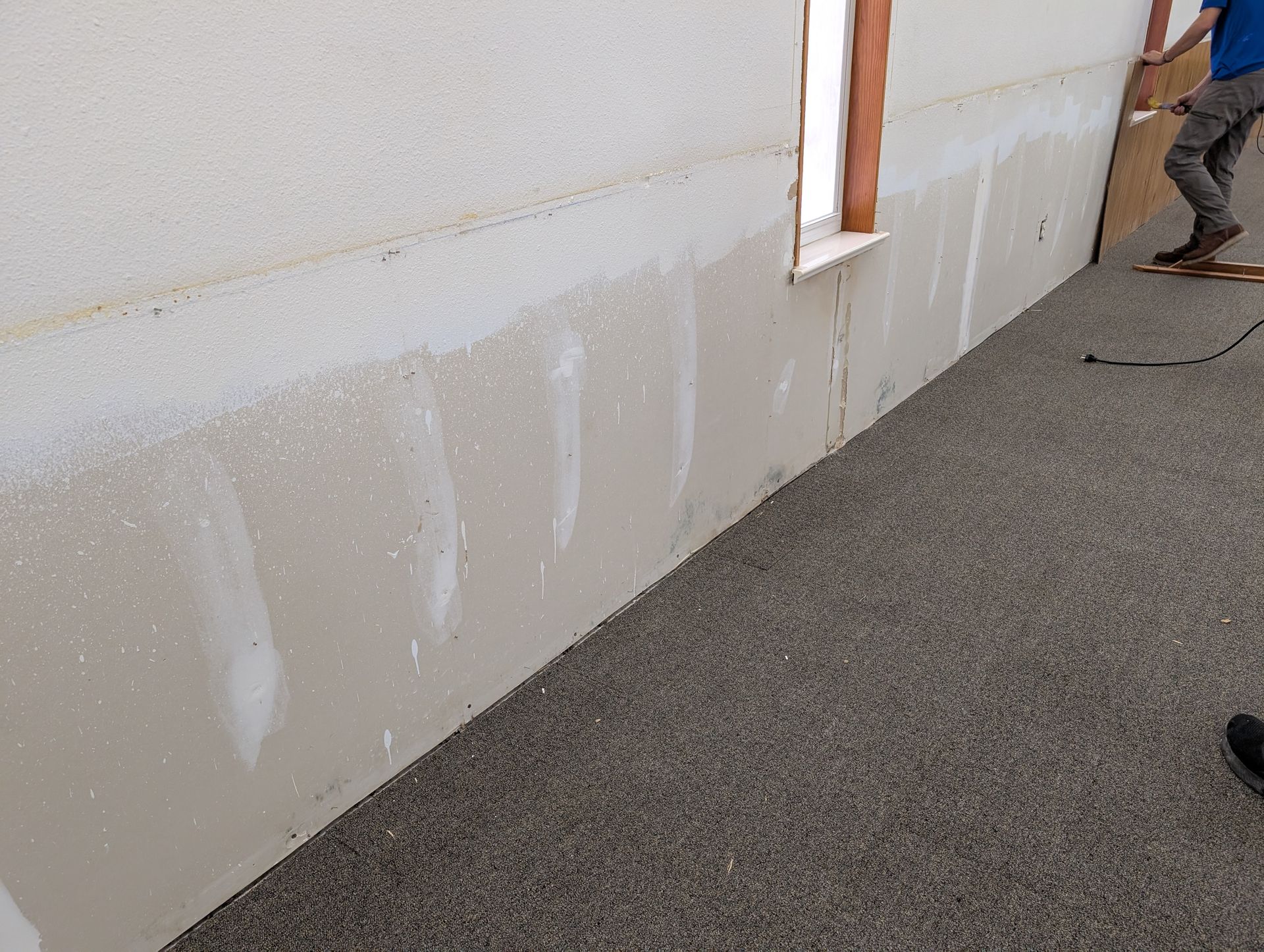 Person installing drywall on a wall with a window; neutral color palette and carpeted floor.