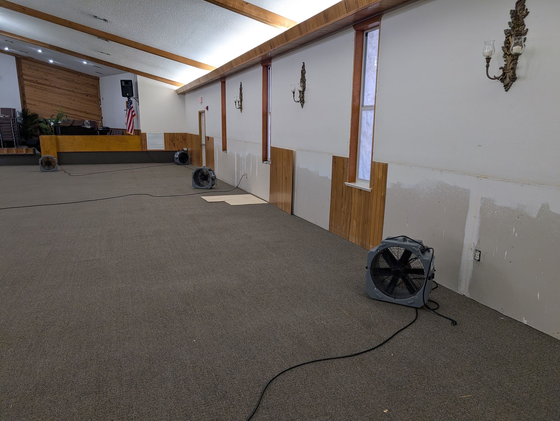 Interior of a room with exposed drywall, fans, and a carpeted floor, likely due to water damage.