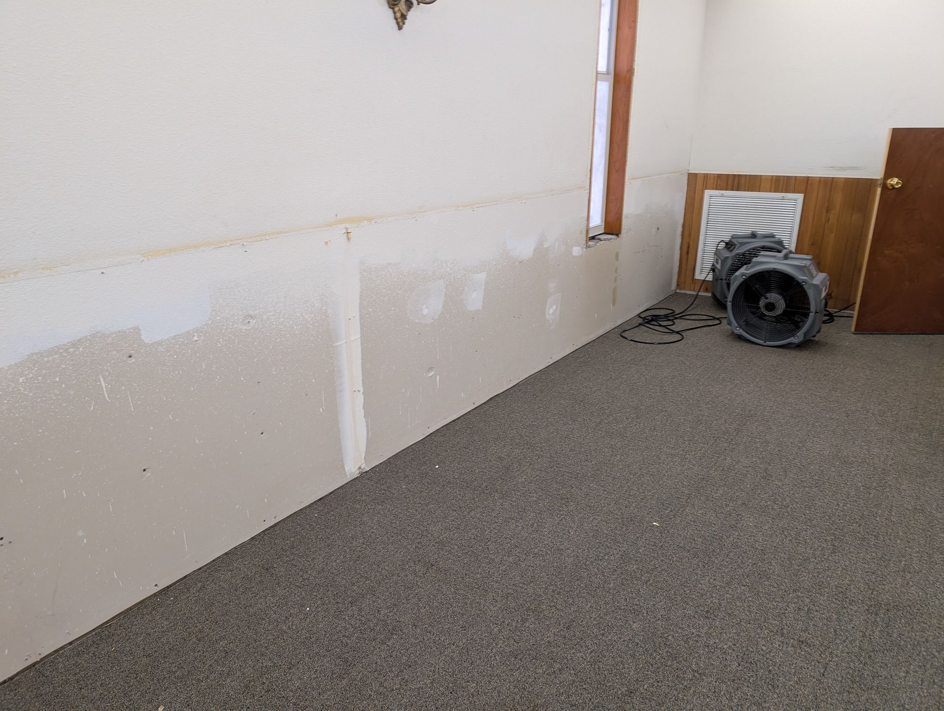 Room with damaged drywall, carpeted floor, window, door, and equipment leaning against a wall.