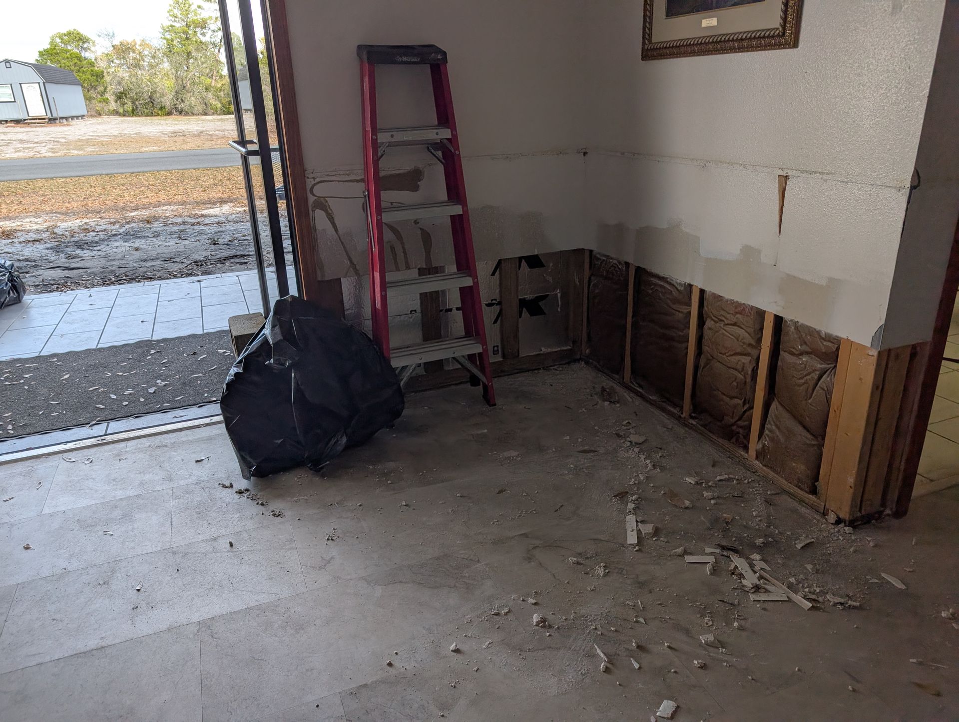 Garage interior with drywall and insulation exposed, ladder, debris, and open garage door.