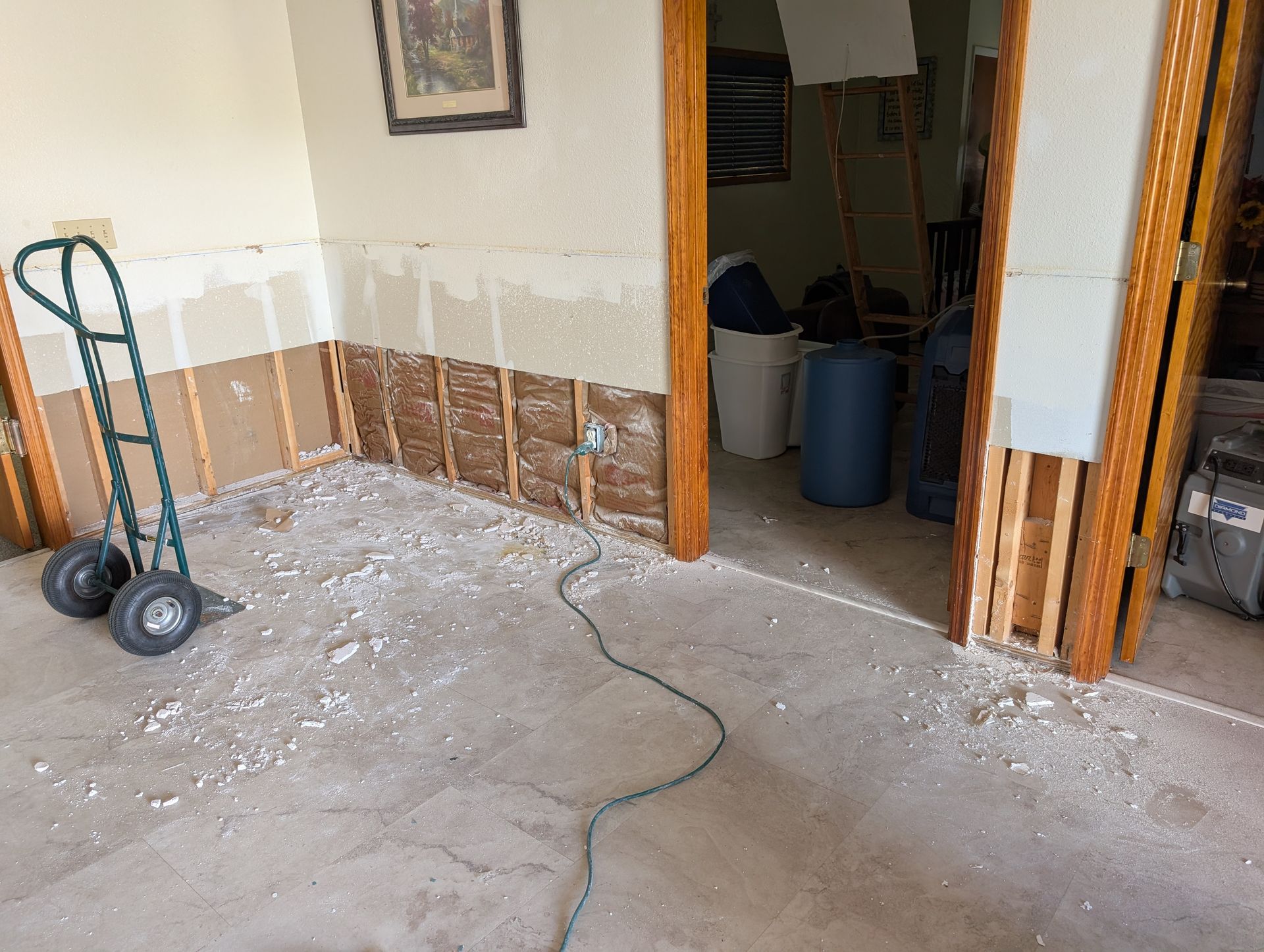 Room with damaged drywall, debris on the floor, hand truck, and open doorway to another room with barrels.