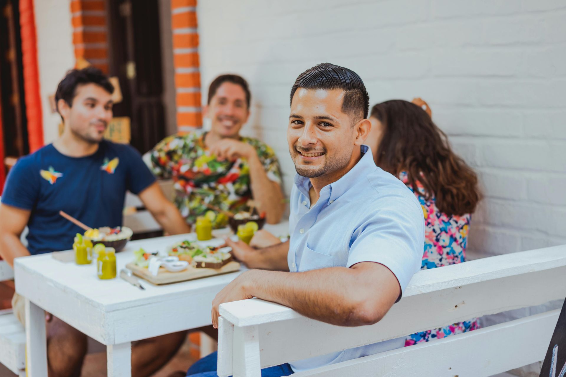 Four people sit at a white table outdoors, eating breakfast and smiling, with a man in the foreground looking at camera.