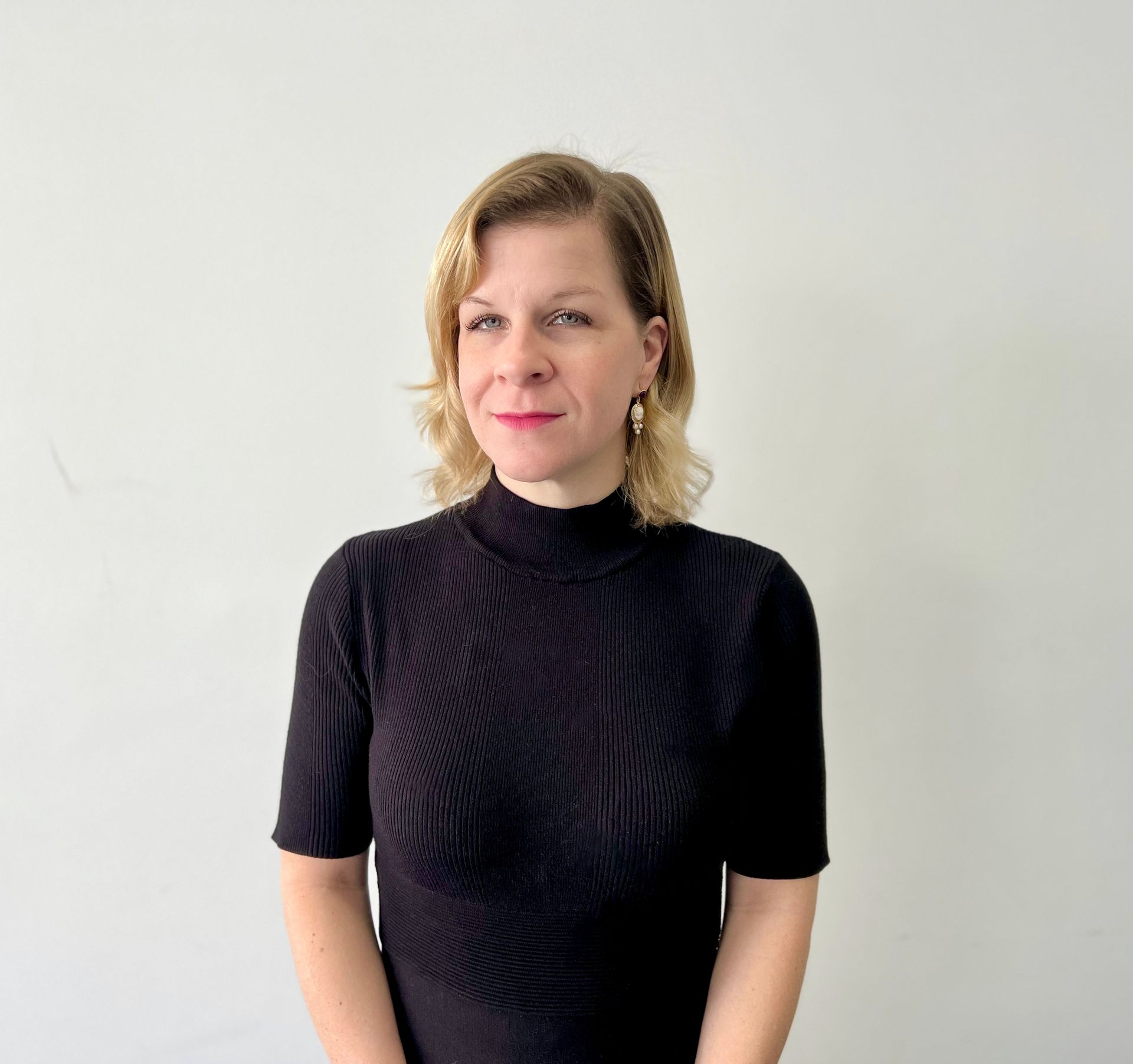 A woman wearing a black sweater and earrings is standing in front of a white wall.