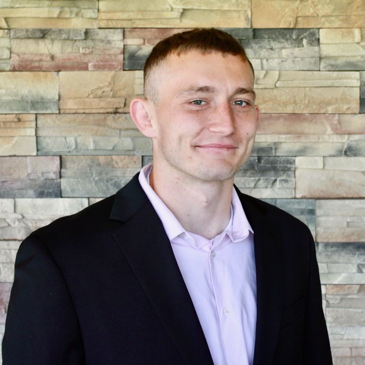 A man in a suit and a purple shirt is smiling in front of a brick wall.