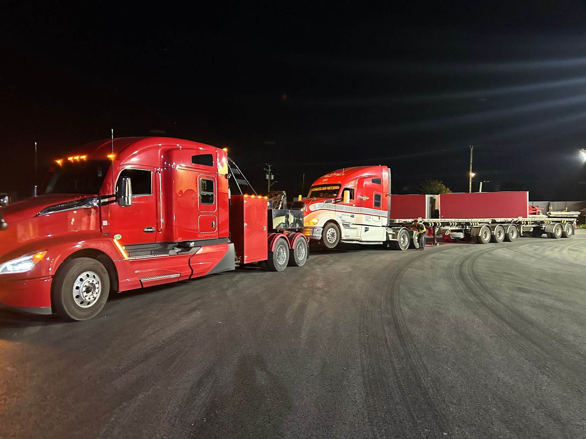 Une dépanneuse rouge tractant un semi-remorque rouge et une remorque la nuit sur un terrain pavé.