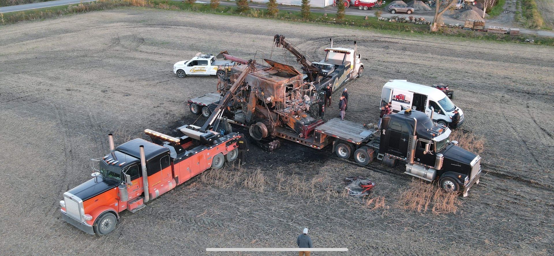 Vue aérienne de plusieurs dépanneuses entourant un semi-remorque gravement endommagé. La scène se déroule dans un champ où se trouvent les décombres d'un incendie.