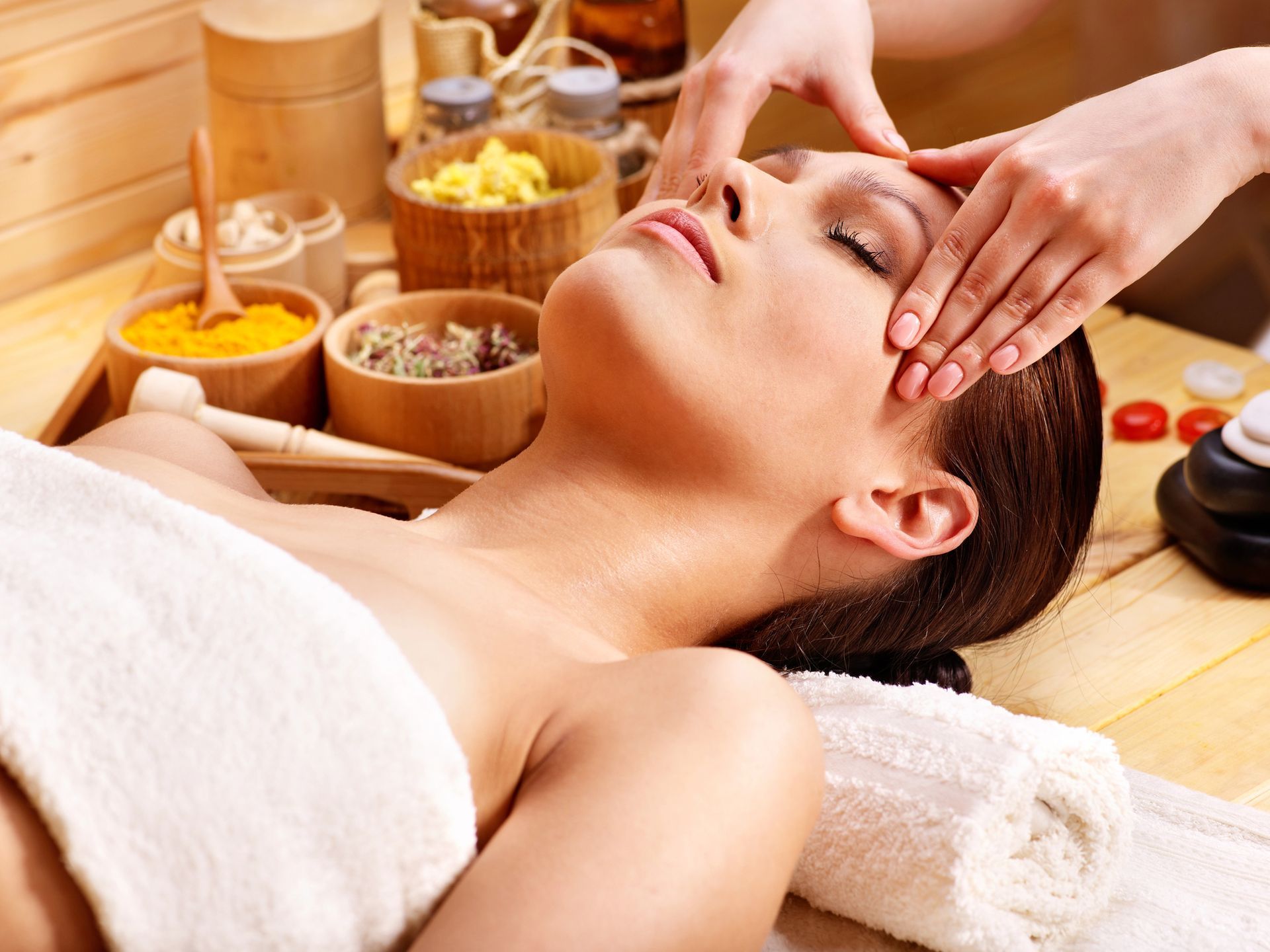 Una terapista di una spa esegue un massaggio facciale su una donna sdraiata su un asciugamano, con candele e ciotole nelle vicinanze.