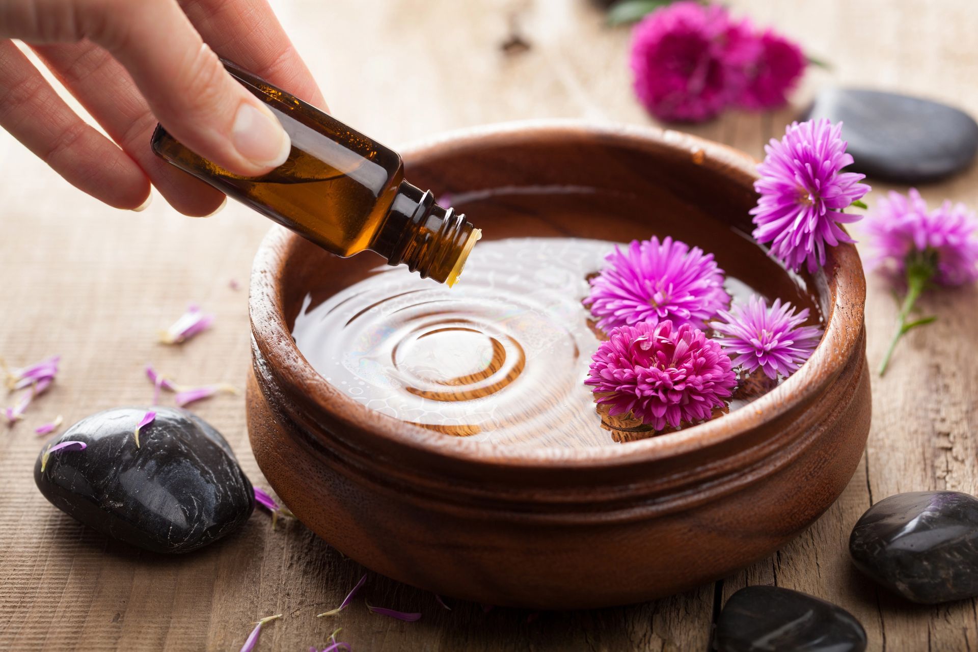 Versare a mano l'olio in una ciotola da spa con fiori rosa e pietre nere su un tavolo di legno.