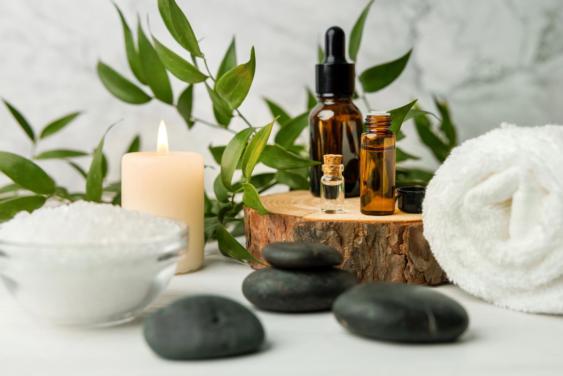 Allestimento da spa con boccette di olio color ambra, candela, pietre, asciugamano, ciotola e foglie verdi su una superficie bianca.