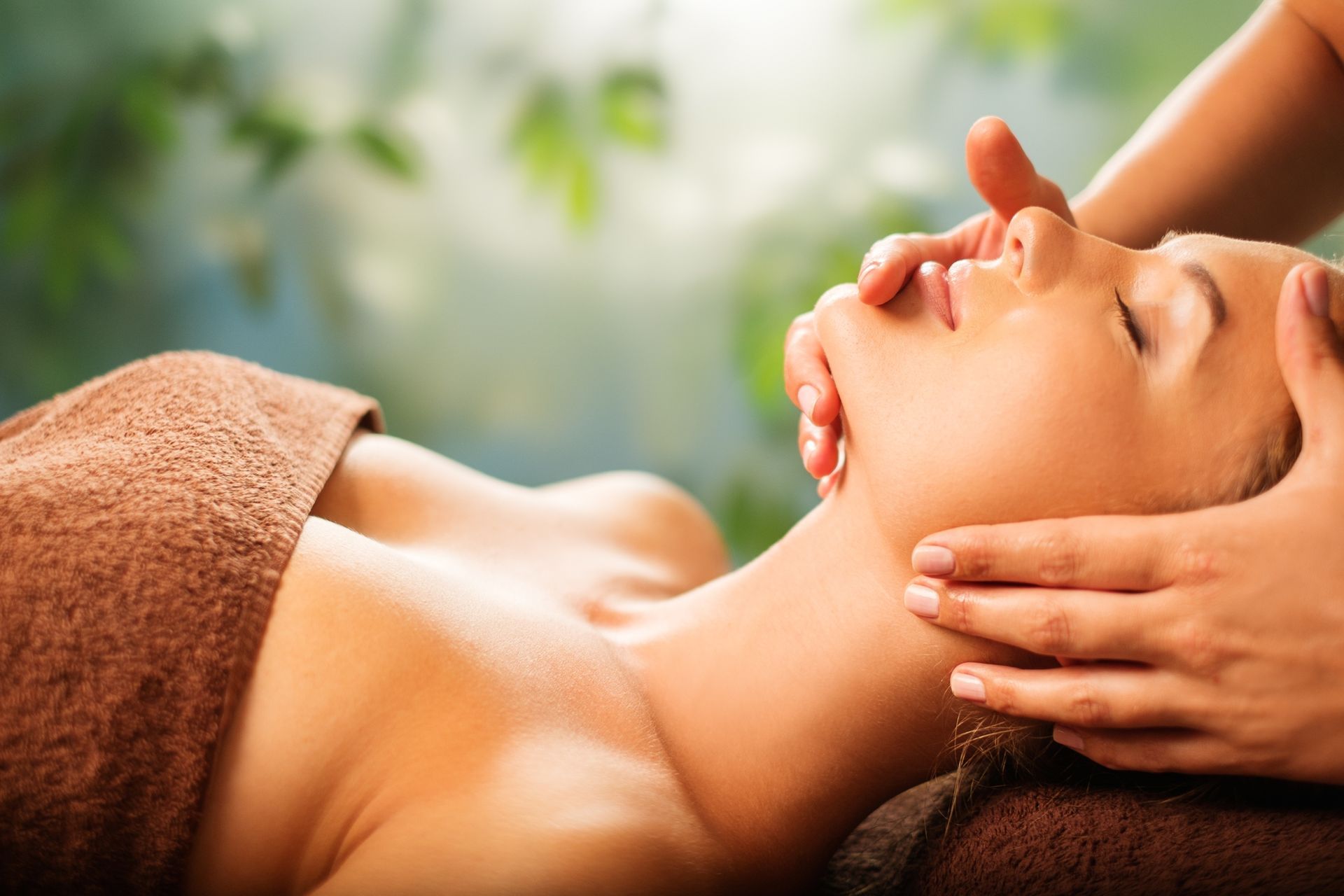 Massaggio in una spa con una persona che riceve un trattamento viso su un asciugamano marrone, mani che accarezzano delicatamente il viso.