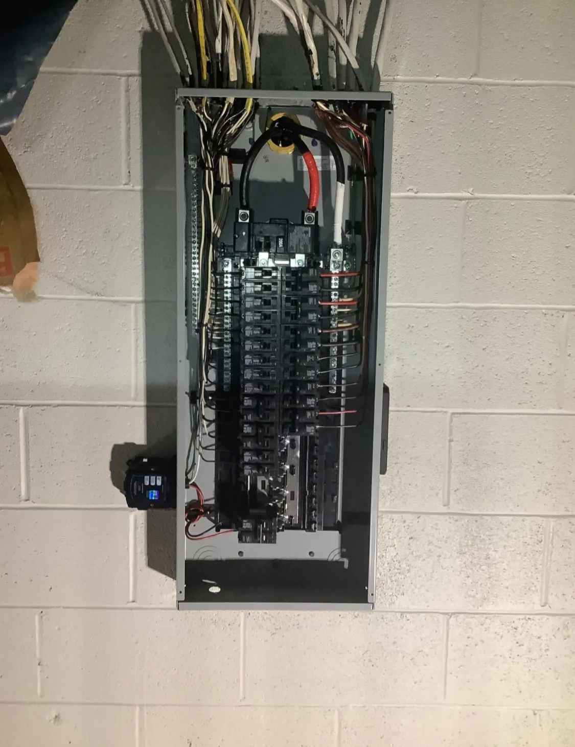 Electrical panel with numerous circuit breakers mounted on a white cinder block wall. Wires are visible above the panel.