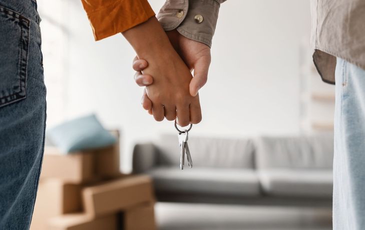 Couple holding hands with keys, standing in a room with boxes, blurred sofa in background.
